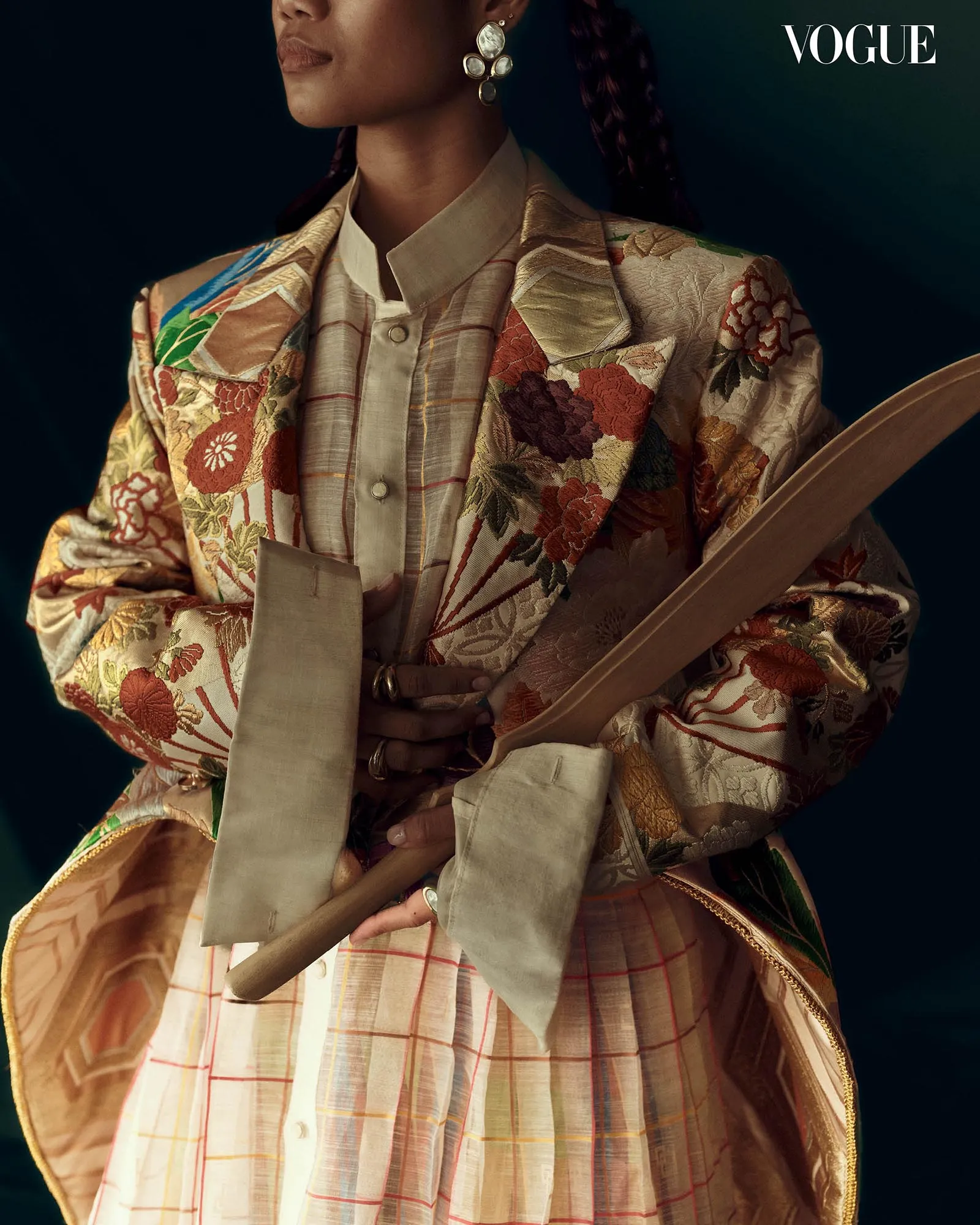 Person wearing an ornate floral brocade blazer over a beige shirt, with hands folded around a wooden prop; Vogue logo in the corner.