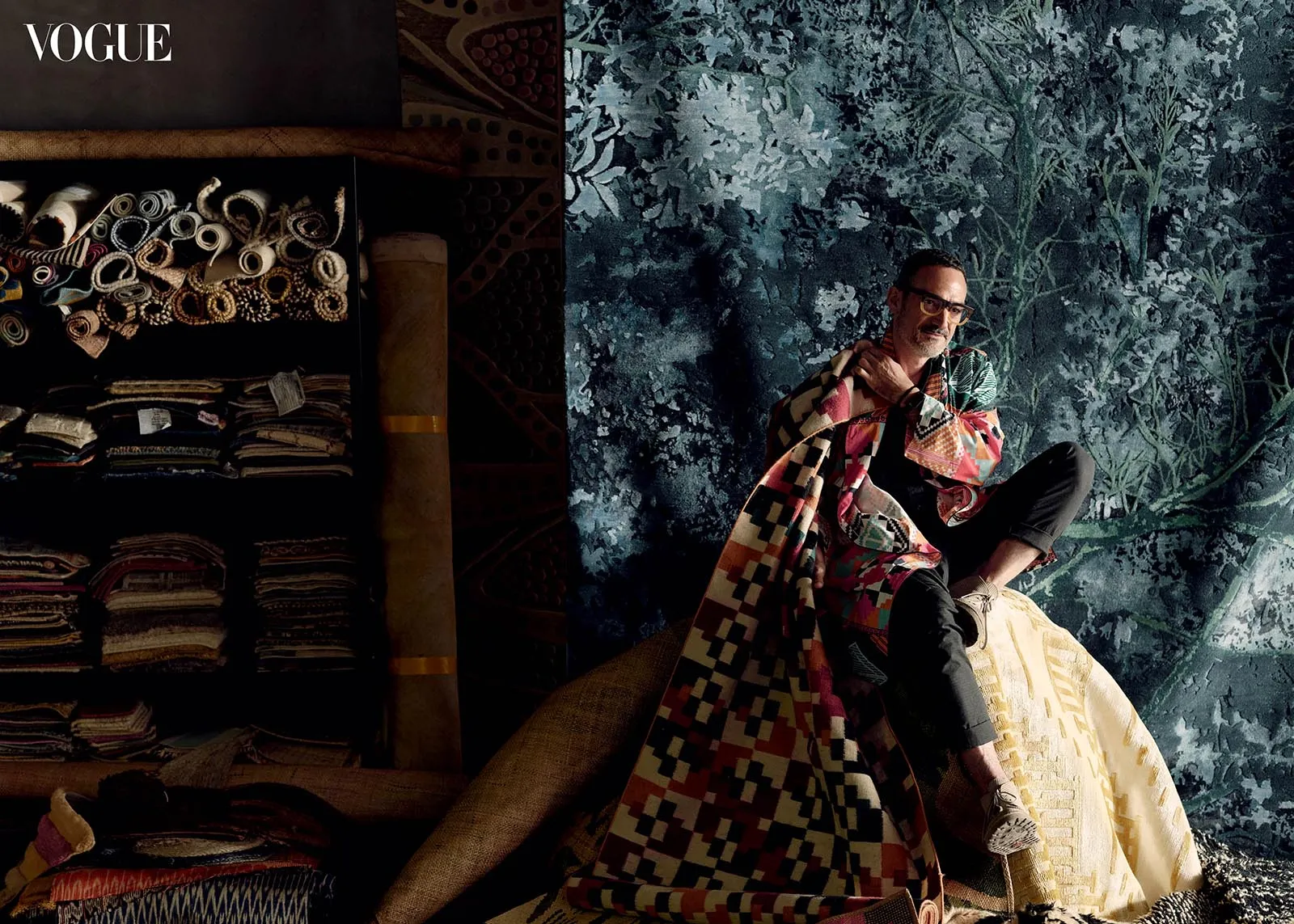 Vogue fashion shoot: man in a colorful patchwork coat sits on a rug against a textured blue botanical backdrop, with textile shelves on the left.