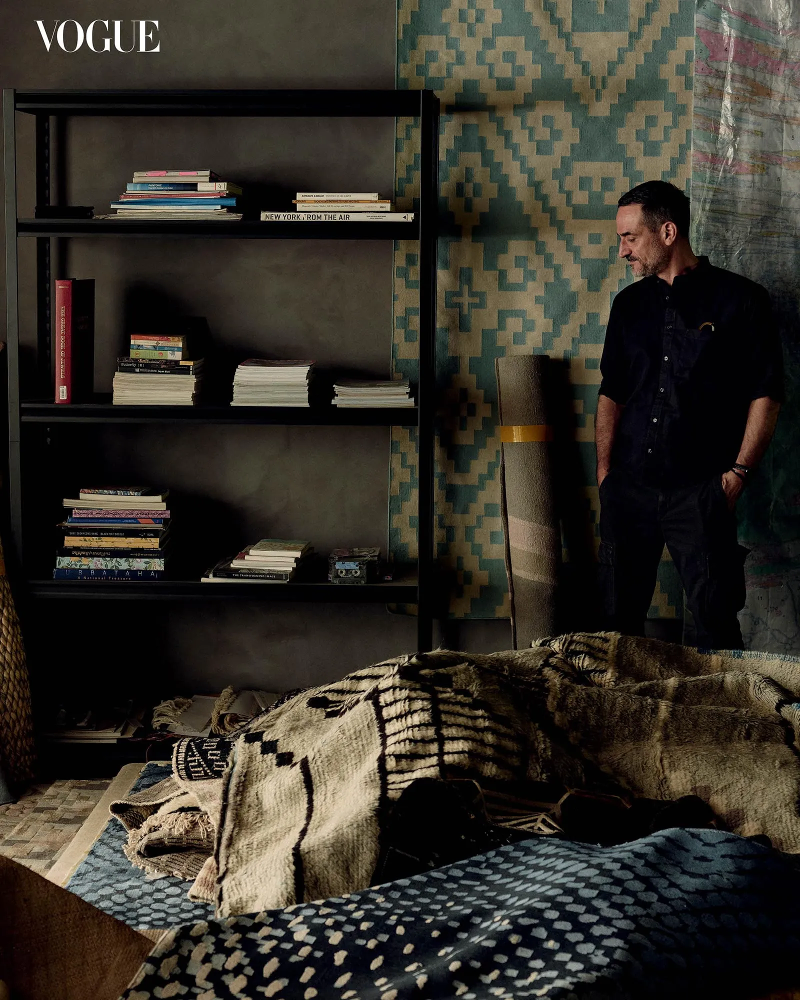 Dimly lit room with a black bookshelf filled with books and magazines; a man in dark clothing leans on a rolled rug beside a teal geometric wall.