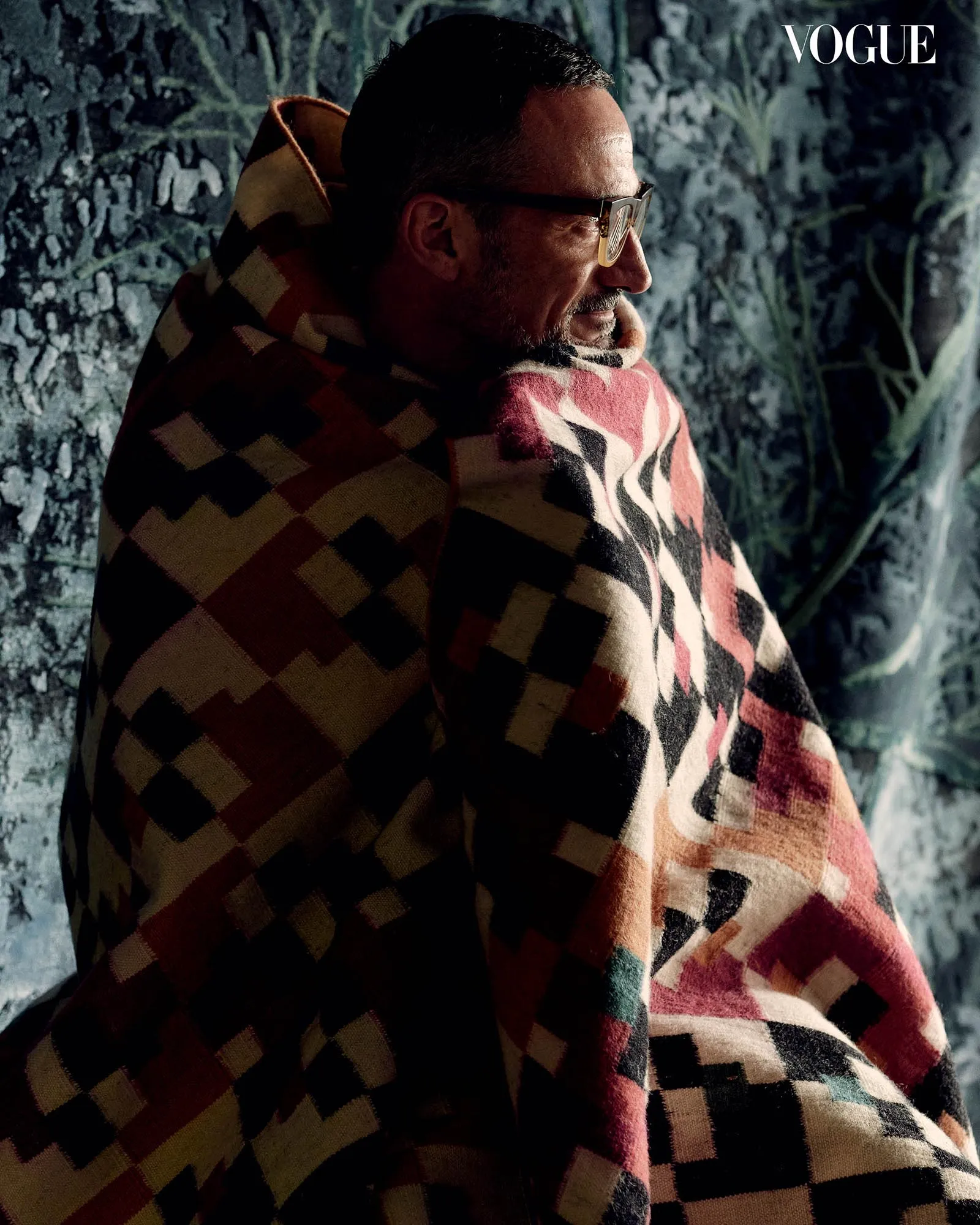 Side profile of a man wearing glasses, wrapped in a red, beige, and black geometric blanket against a textured wall.