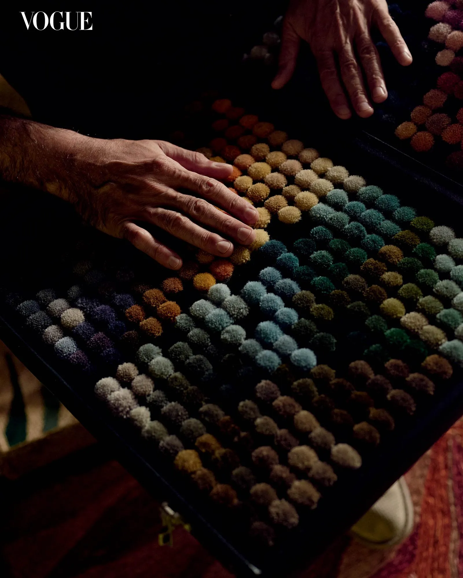 Close-up of hands arranging a rainbow grid of small fuzzy pom-poms on a dark surface, with Vogue visible in the corner.