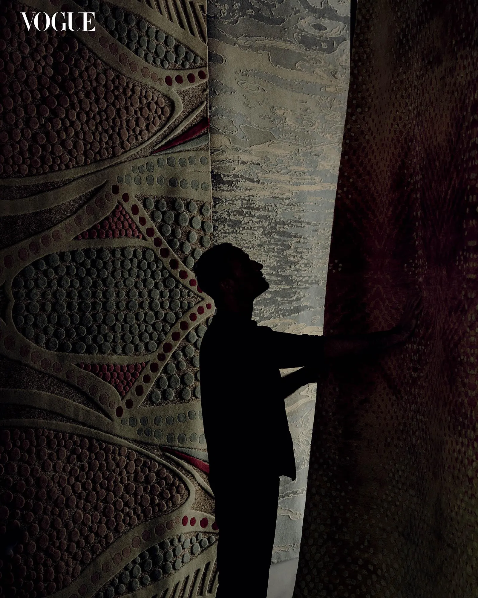 Silhouette of a person standing against a large, dotted wall mural with the Vogue logo in the top-left corner.