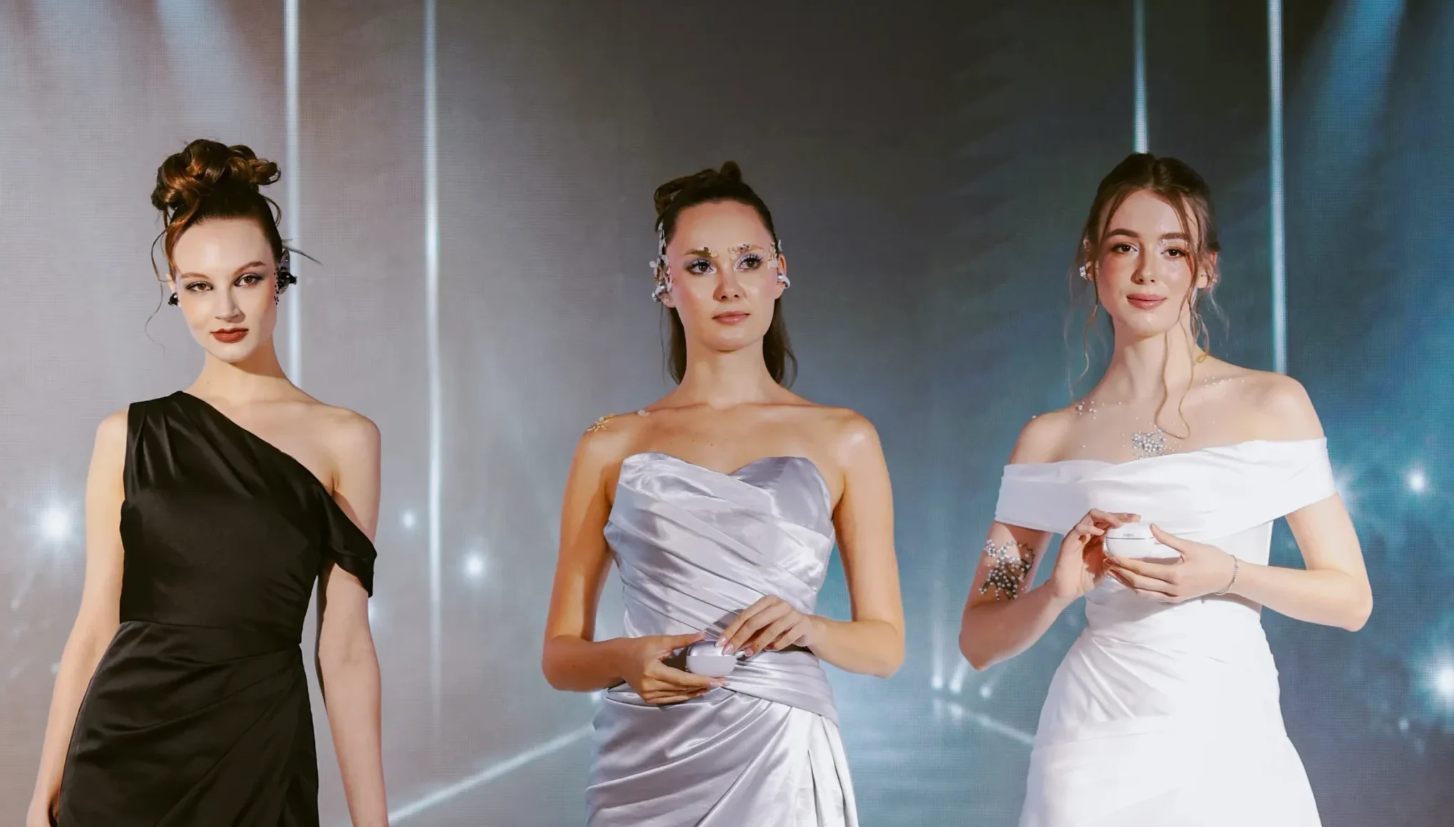 Three models in evening gowns posing on a lit runway stage with spotlights overhead (black, silver, white gowns).
