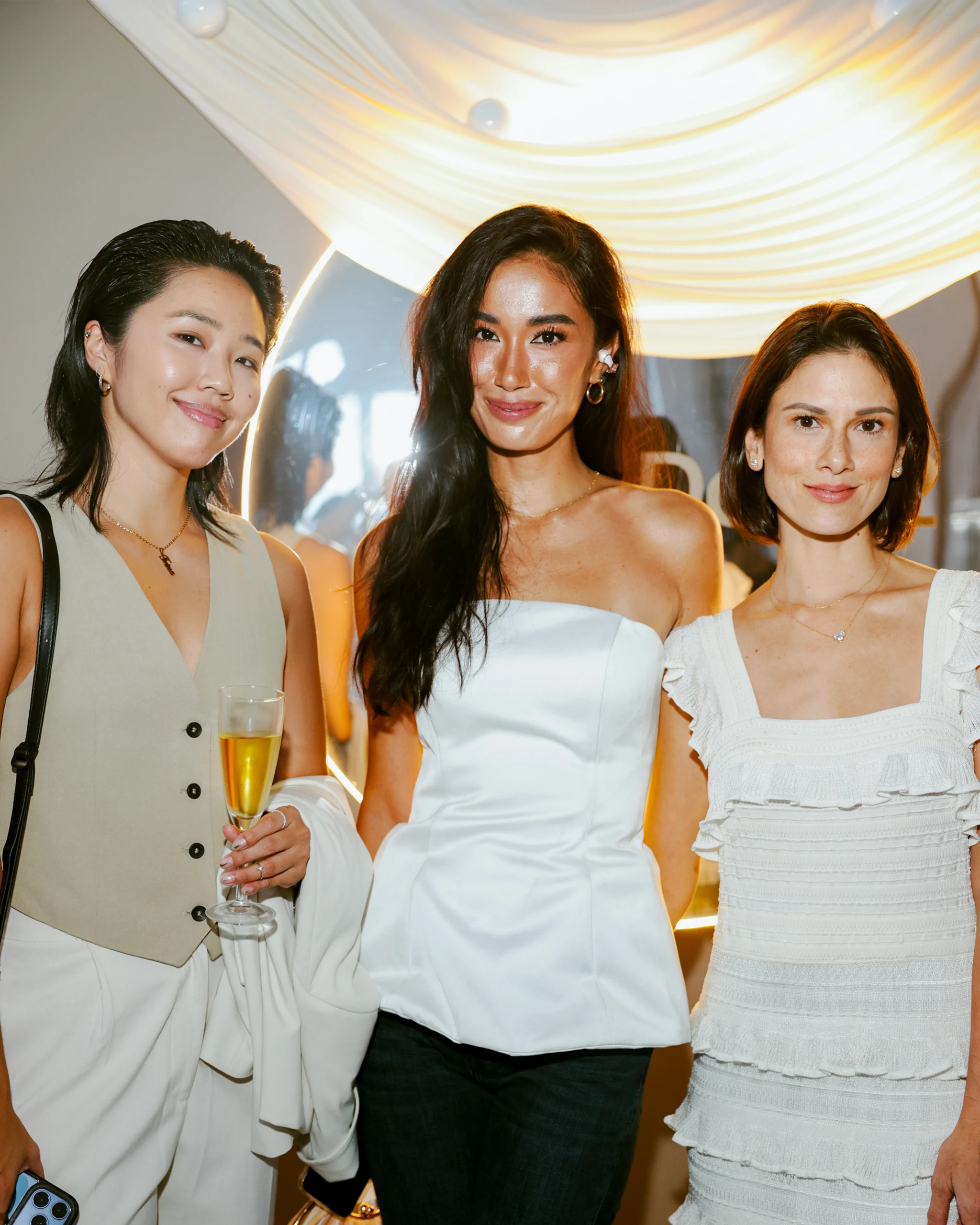Three women posing together at a party, each smiling; one holds a flute of champagne, draped lights in the background.