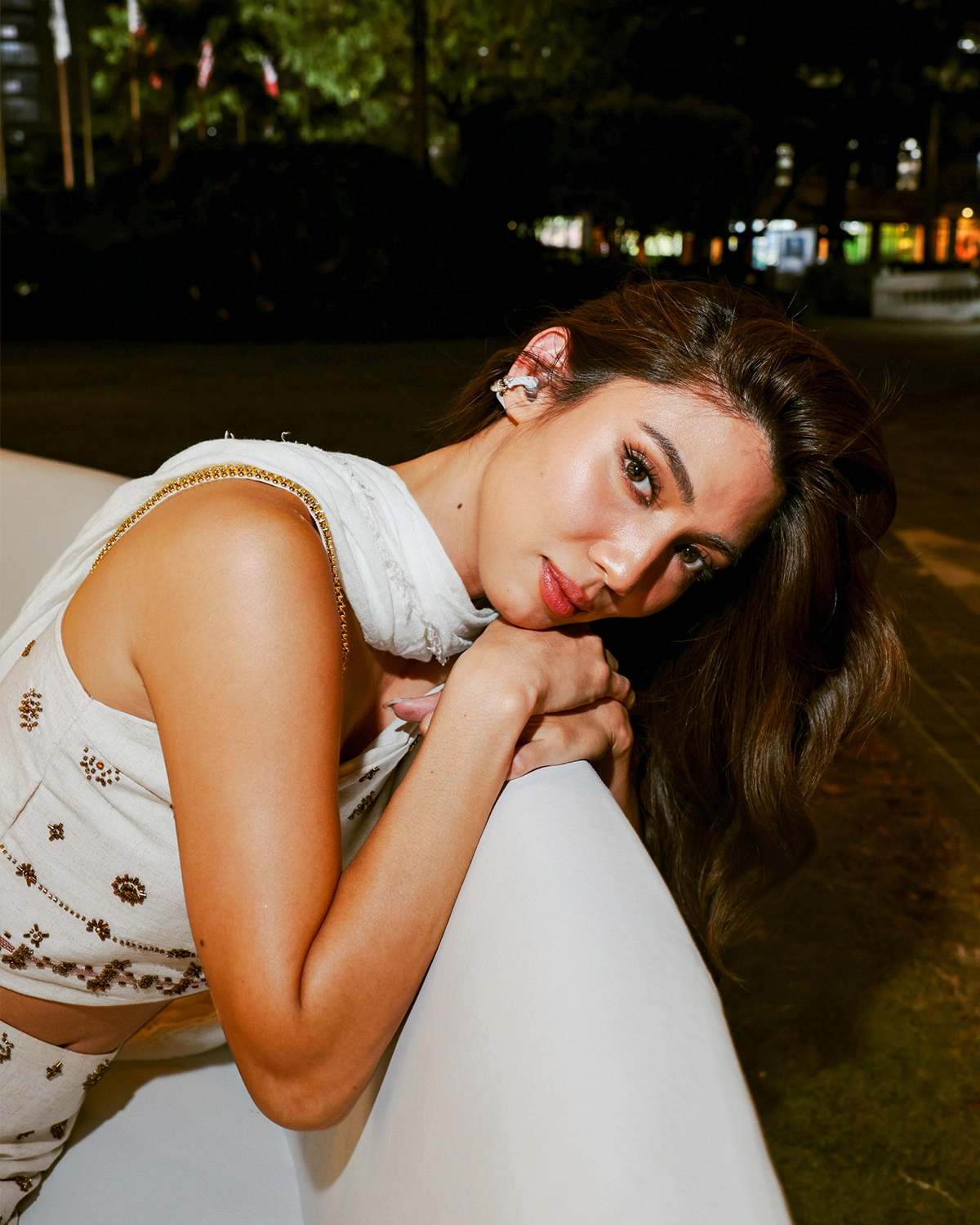 Woman with long brown hair leaning on a white railing at night, wearing a white embroidered outfit and scarf in an outdoor setting.