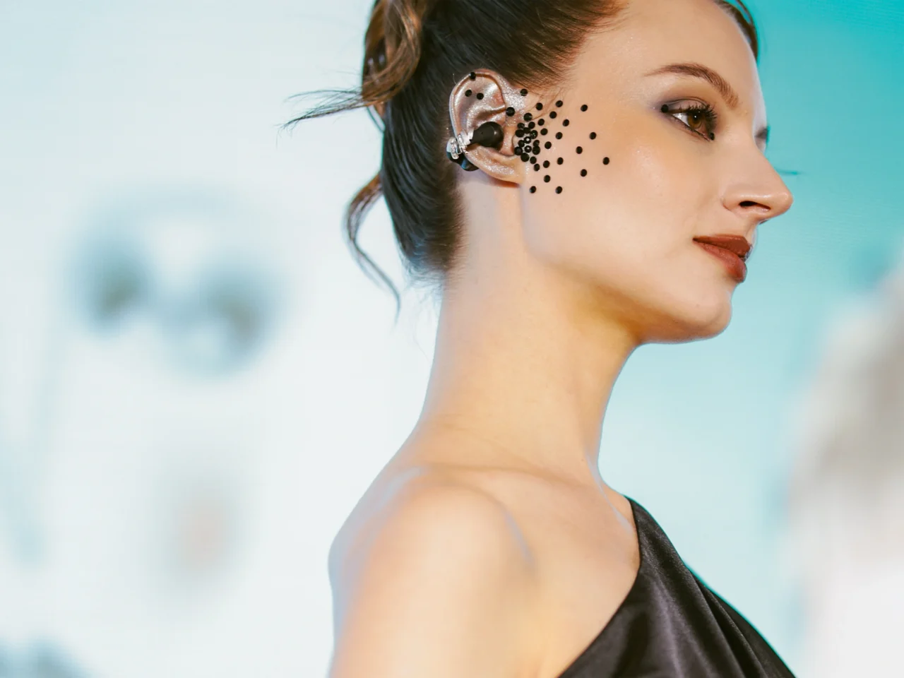 Side profile of a woman wearing an in-ear monitor; black circular dots decorate her cheek near the ear.