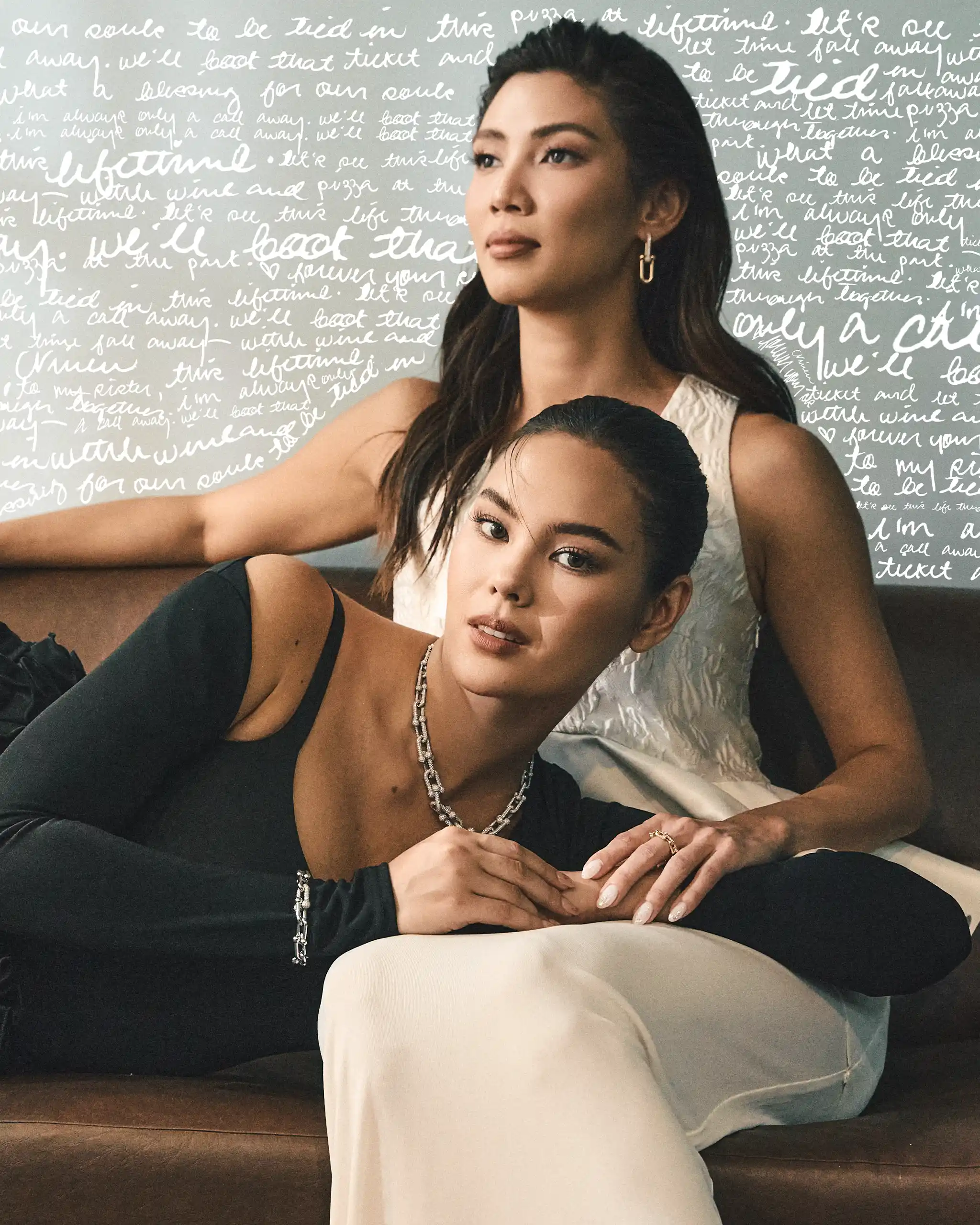 Two women sit close on a sofa; one wears black with a silver chain, the other wears white, with a backdrop of white handwritten script.