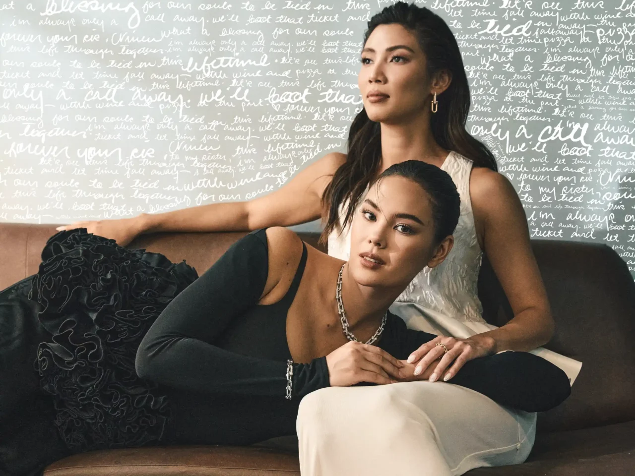 Two women pose together on a sofa against a pale backdrop with white cursive writing; one in a black dress, the other in a light top.
