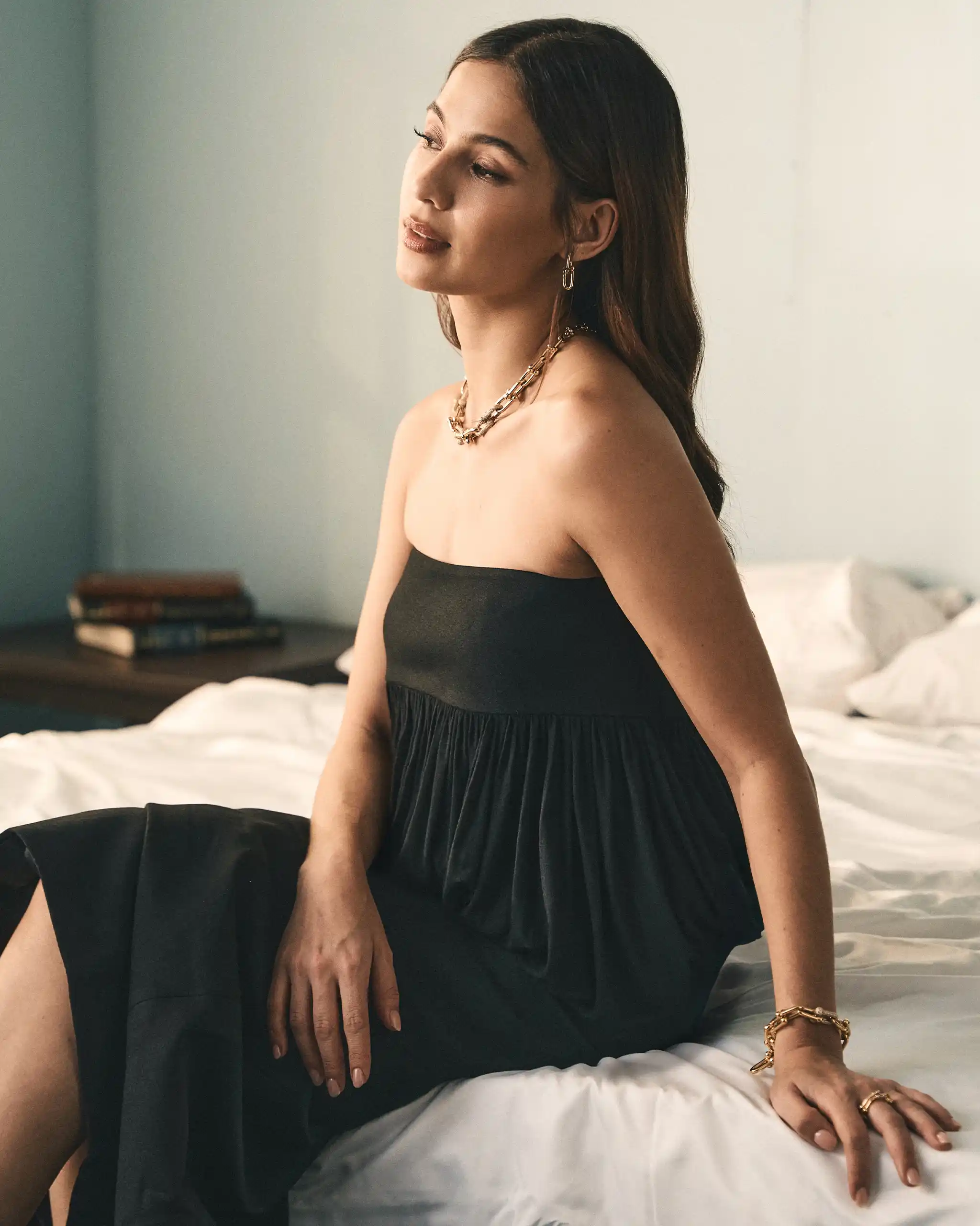 Woman in a strapless black dress sits on a bed, facing left, with gold jewelry and a calm expression.