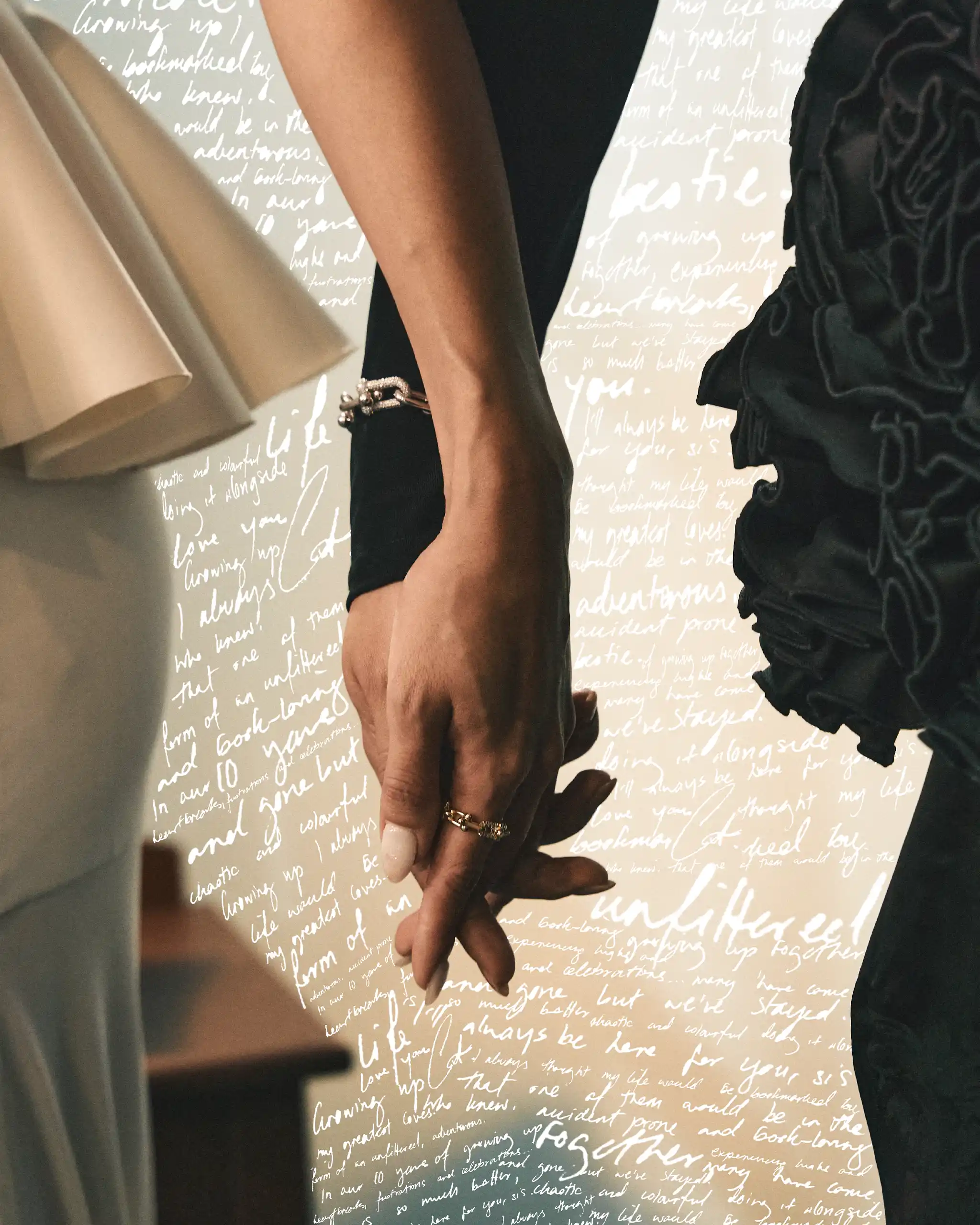 Close-up of two people standing together, one wearing a beige dress and the other a black outfit, with their hands visible and bracelets; handwritten background text behind them.