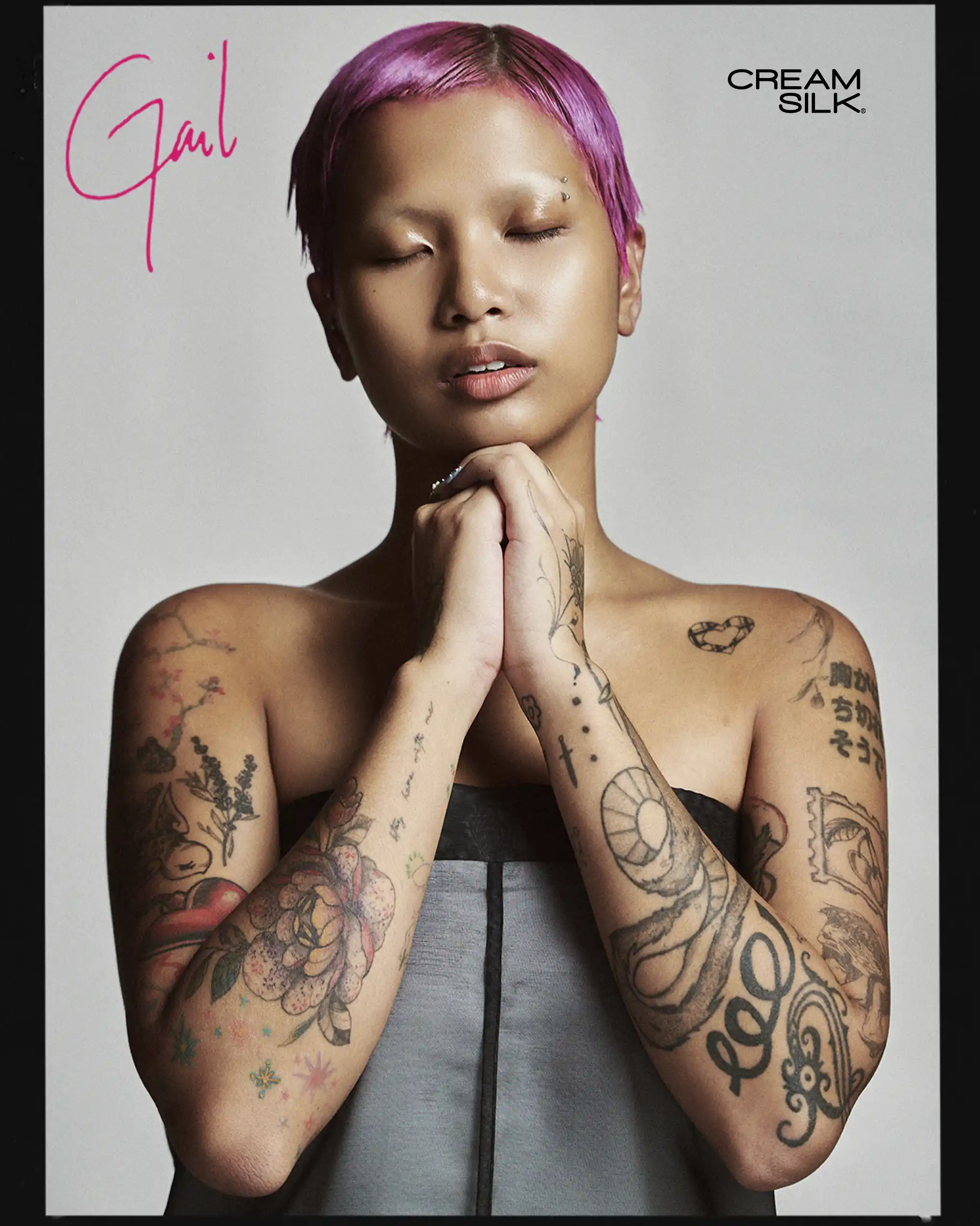 Portrait of a tattooed person with pink hair and eyes closed, hands clasped under chin, wearing a gray strapless top; Cream Silk logo in top right.