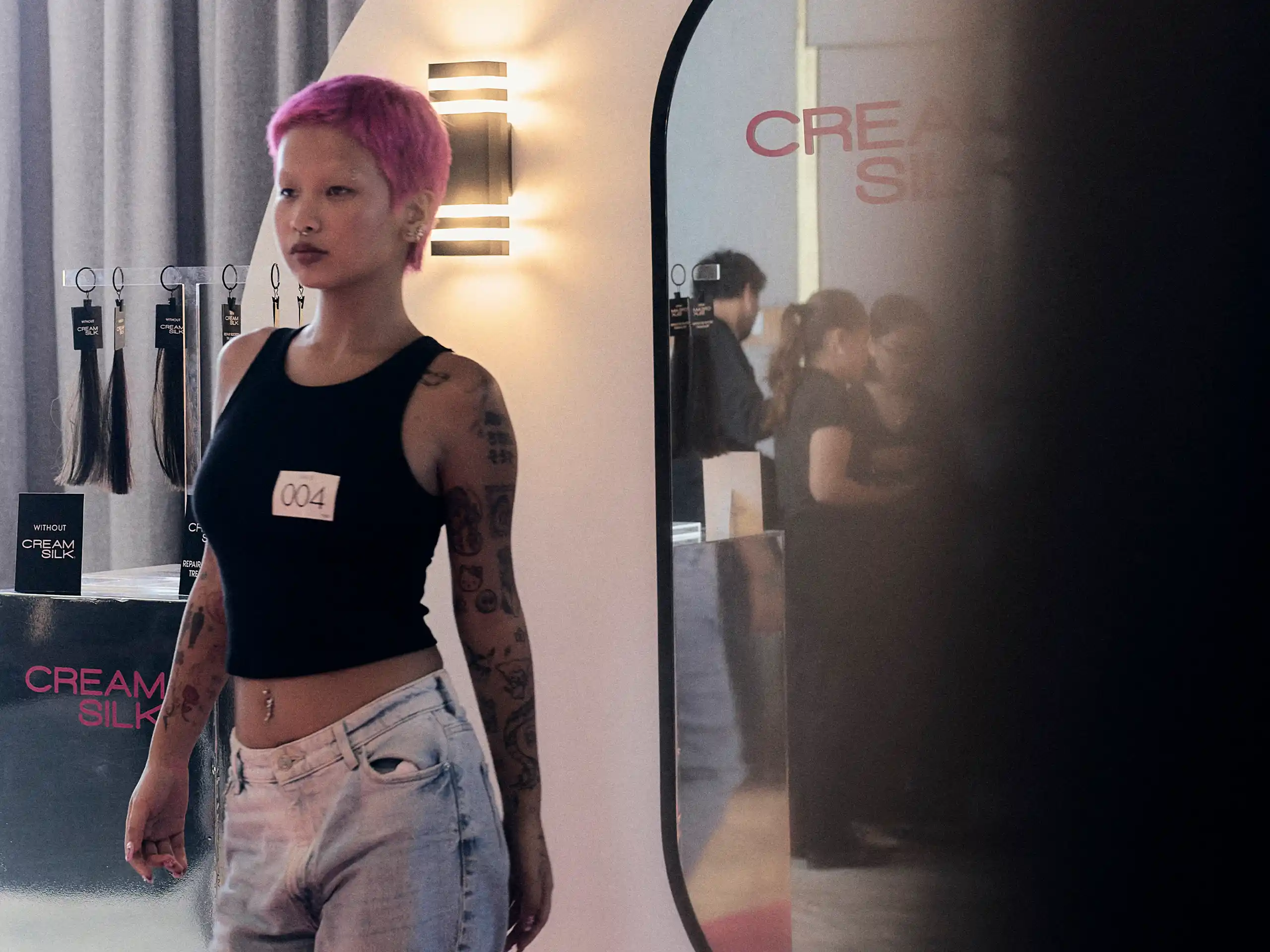 Model with pink hair and tattoos stands beside a hair-extension display labeled 'Cream Silk' in a salon setting.