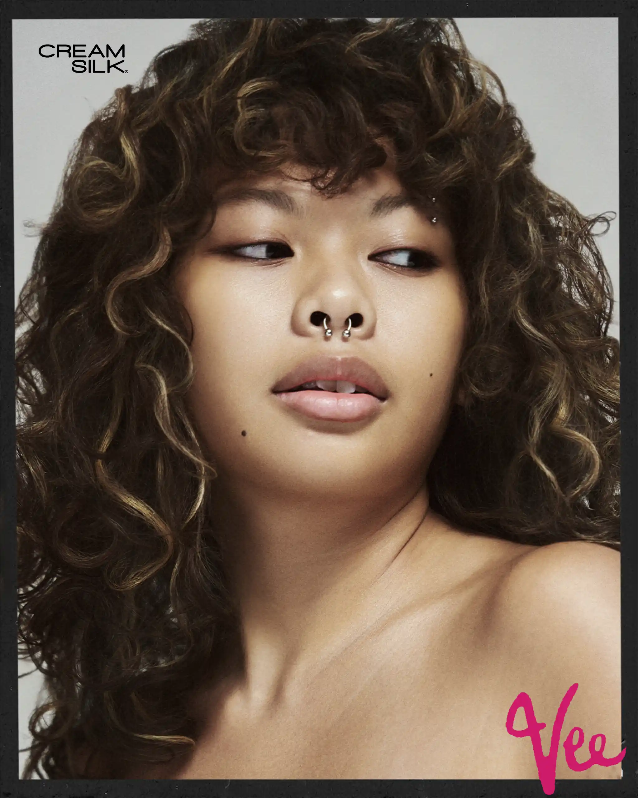 Close-up portrait of a woman with voluminous curly brown hair and a septum piercing, looking to the side.