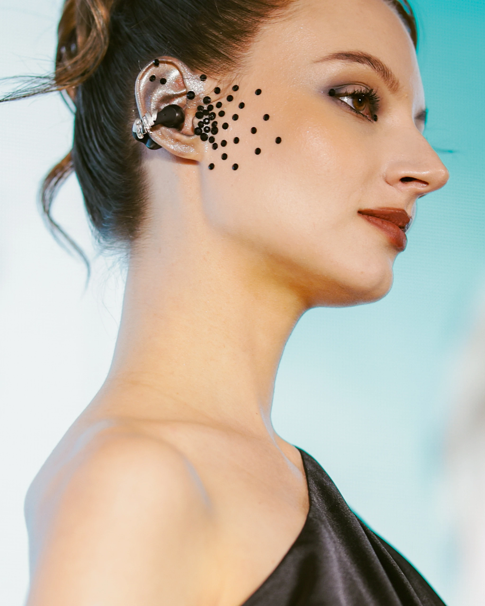 Side profile of a woman wearing an in-ear monitor; black circular dots decorate her cheek near the ear.