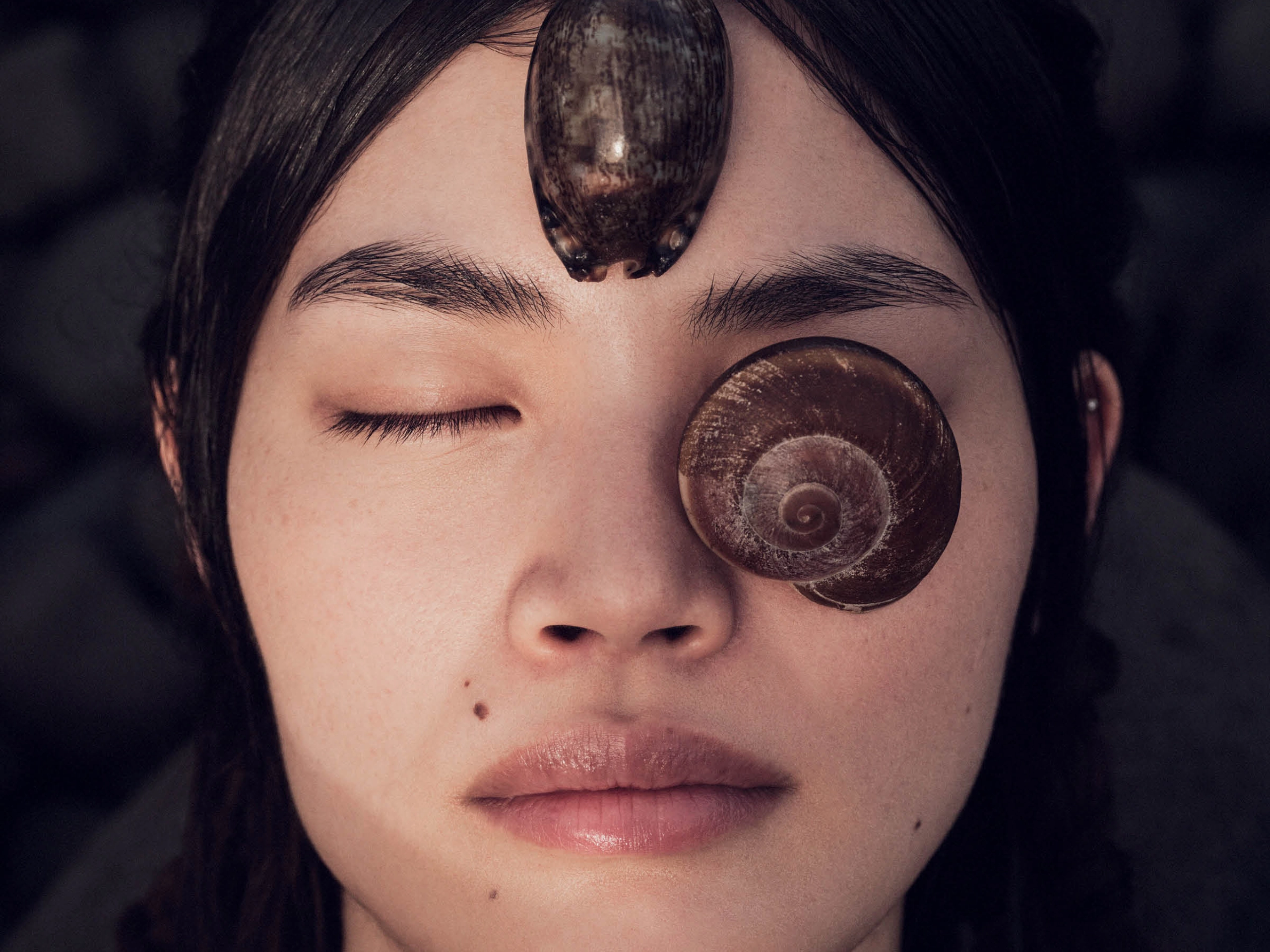 Close-up of a woman's face with a large spiral seashell on her forehead and another shell on her cheek, eyes closed.