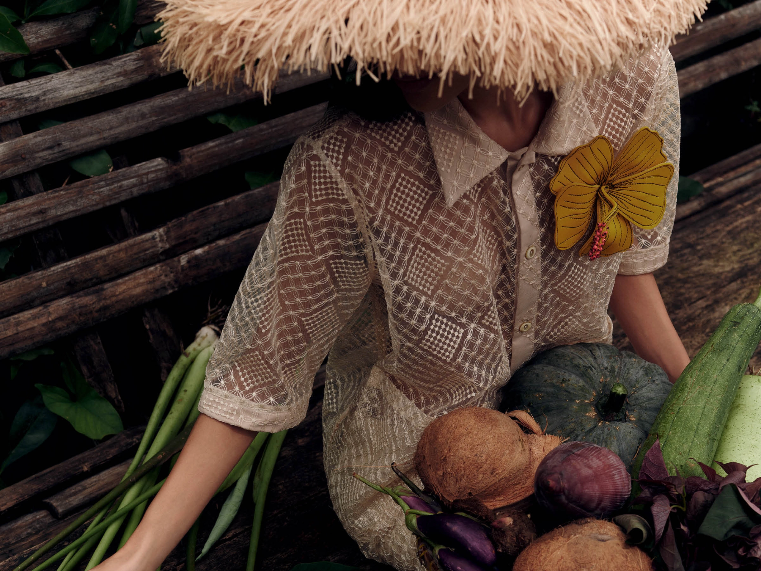 Person in a beige patterned dress and straw hat lies among vegetables on a wooden surface, with a yellow flower brooch.