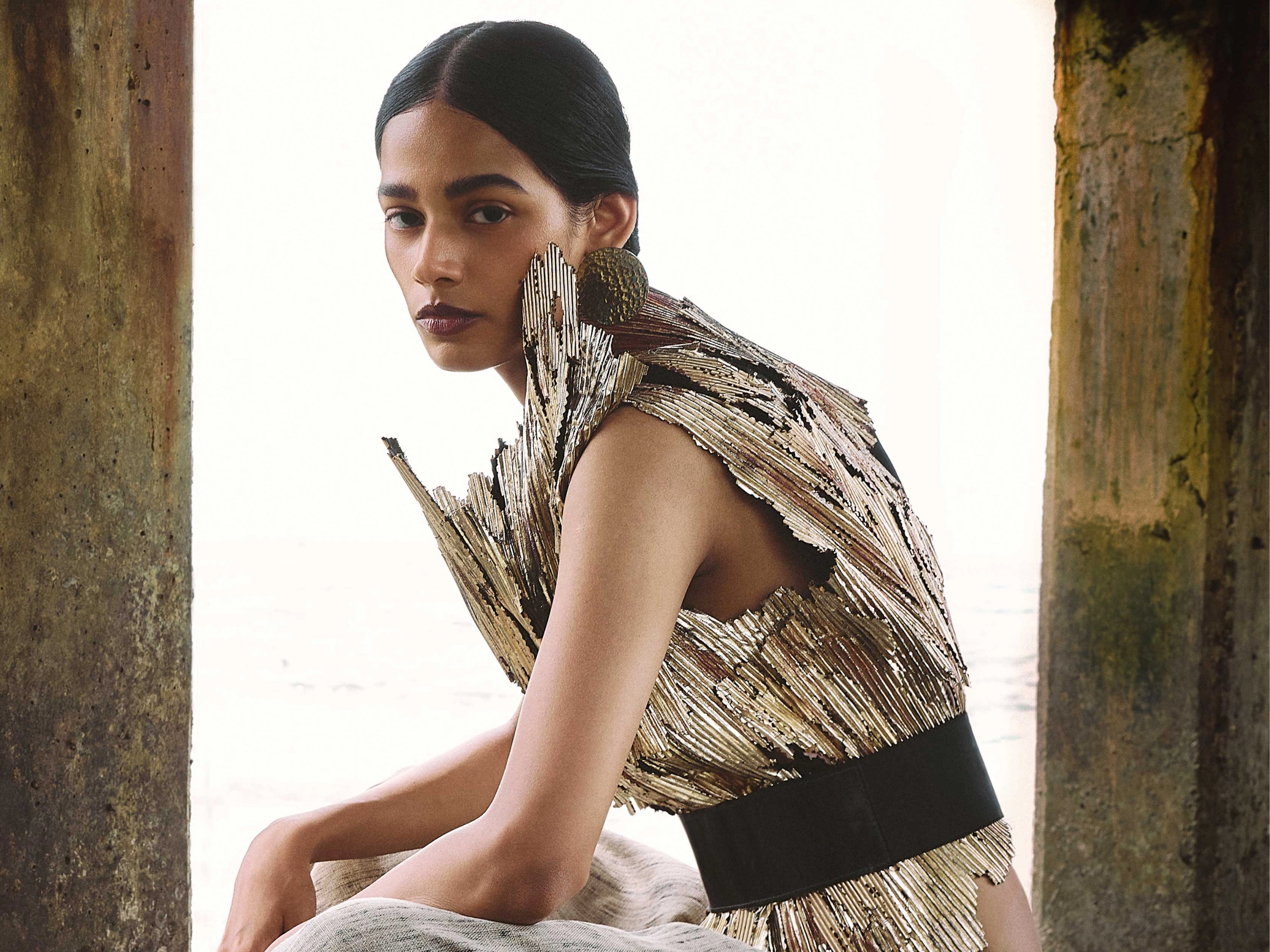 Model wearing a metallic, fringe-textured top sits between weathered columns by the sea. (fashion shoot)