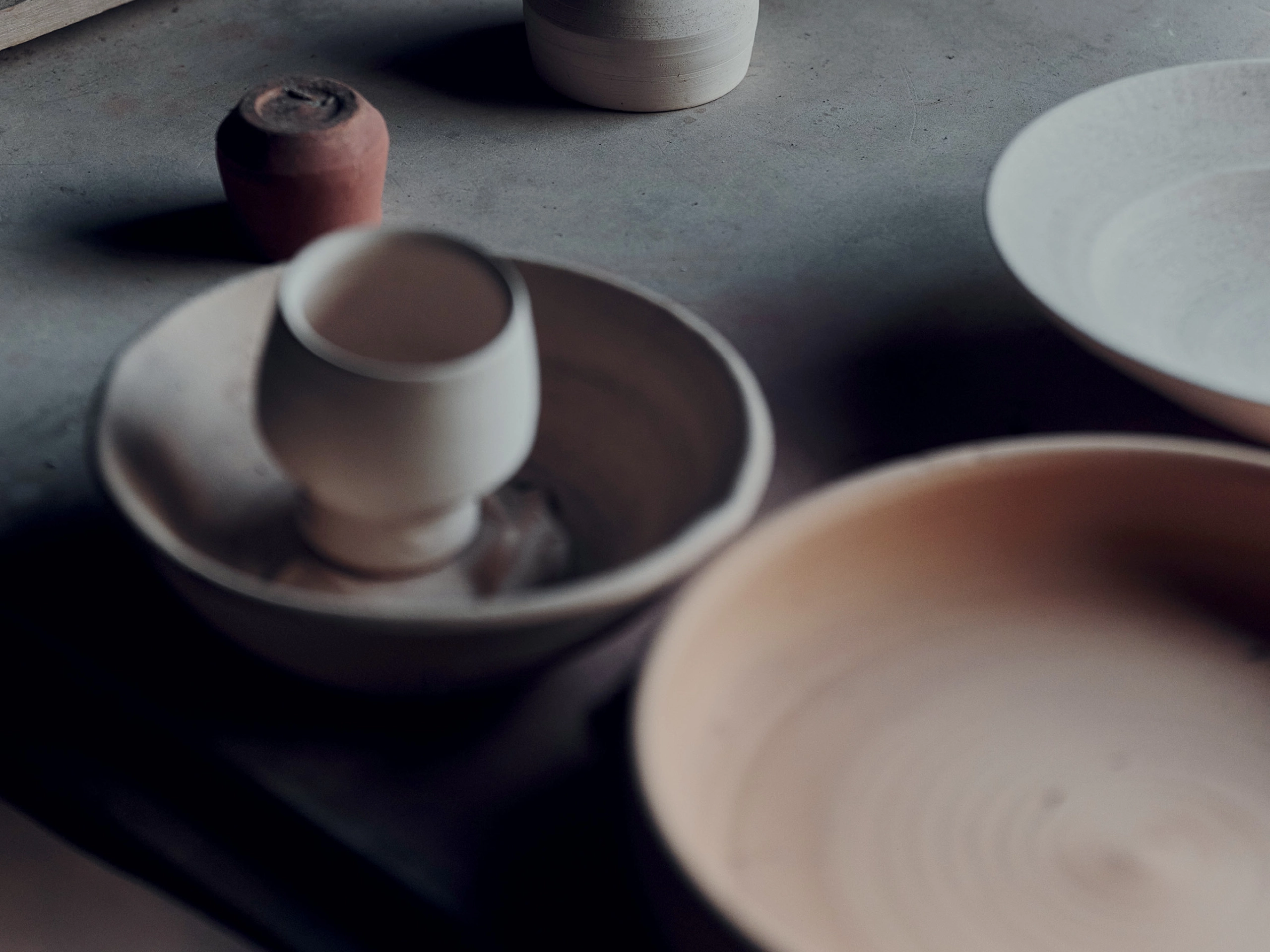 Blurred close-up of ceramic bowls and cups resting on a gray surface or table edge, showing muted earthy tones below the focus point on the left and right.