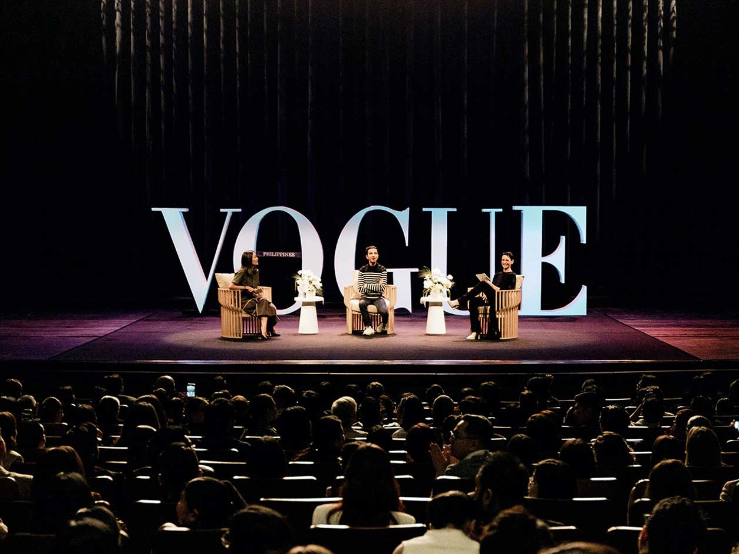 Stage panel on a Vogue backdrop with large letters; four people seated in chairs on stage, audience in foreground.