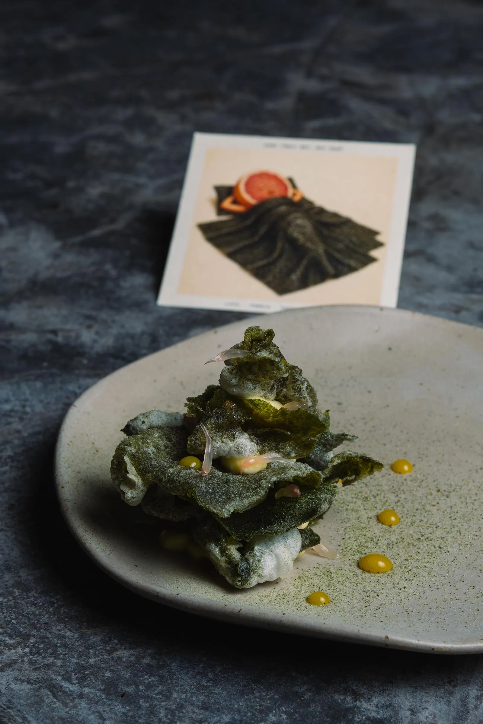 Crispy green tempura stacked on a light plate, with small yellow sauce drops; a blurred card sits in the background.