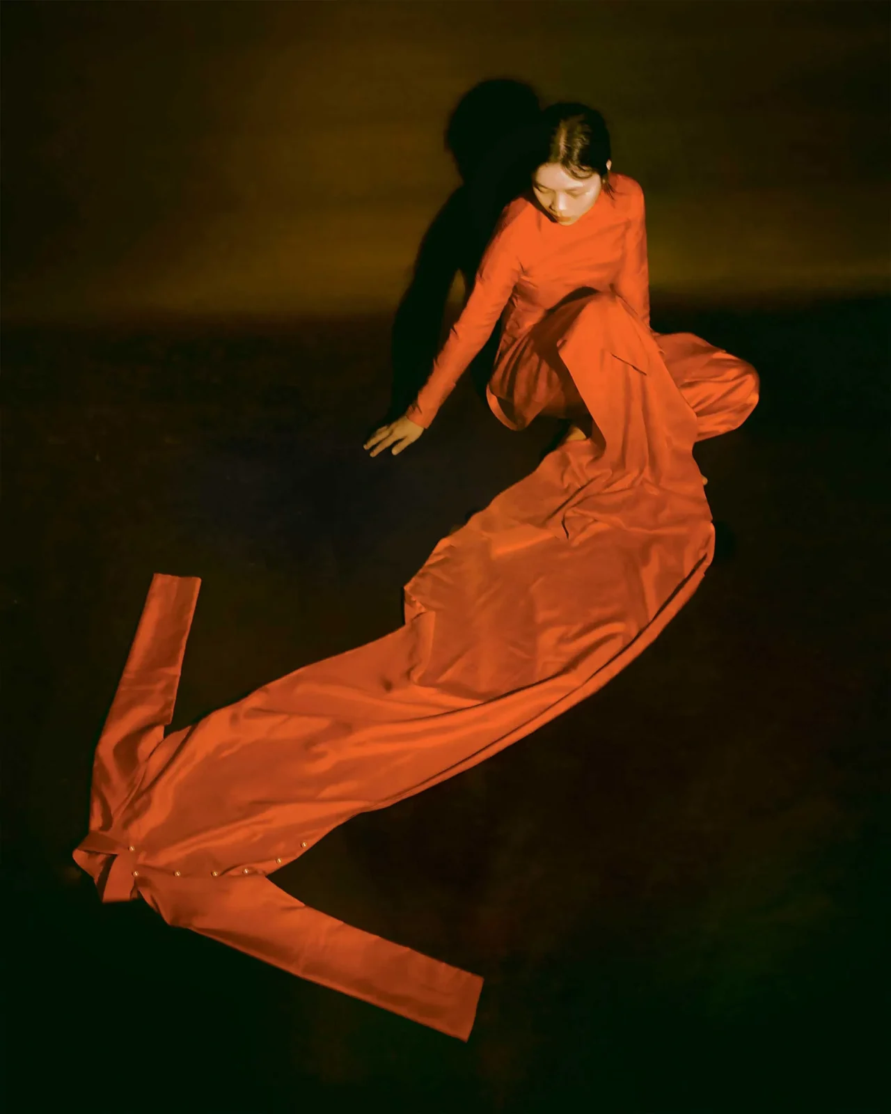 Person in a flowing orange dress crouches on a dark stage bathed in warm orange light, performing a dance pose.