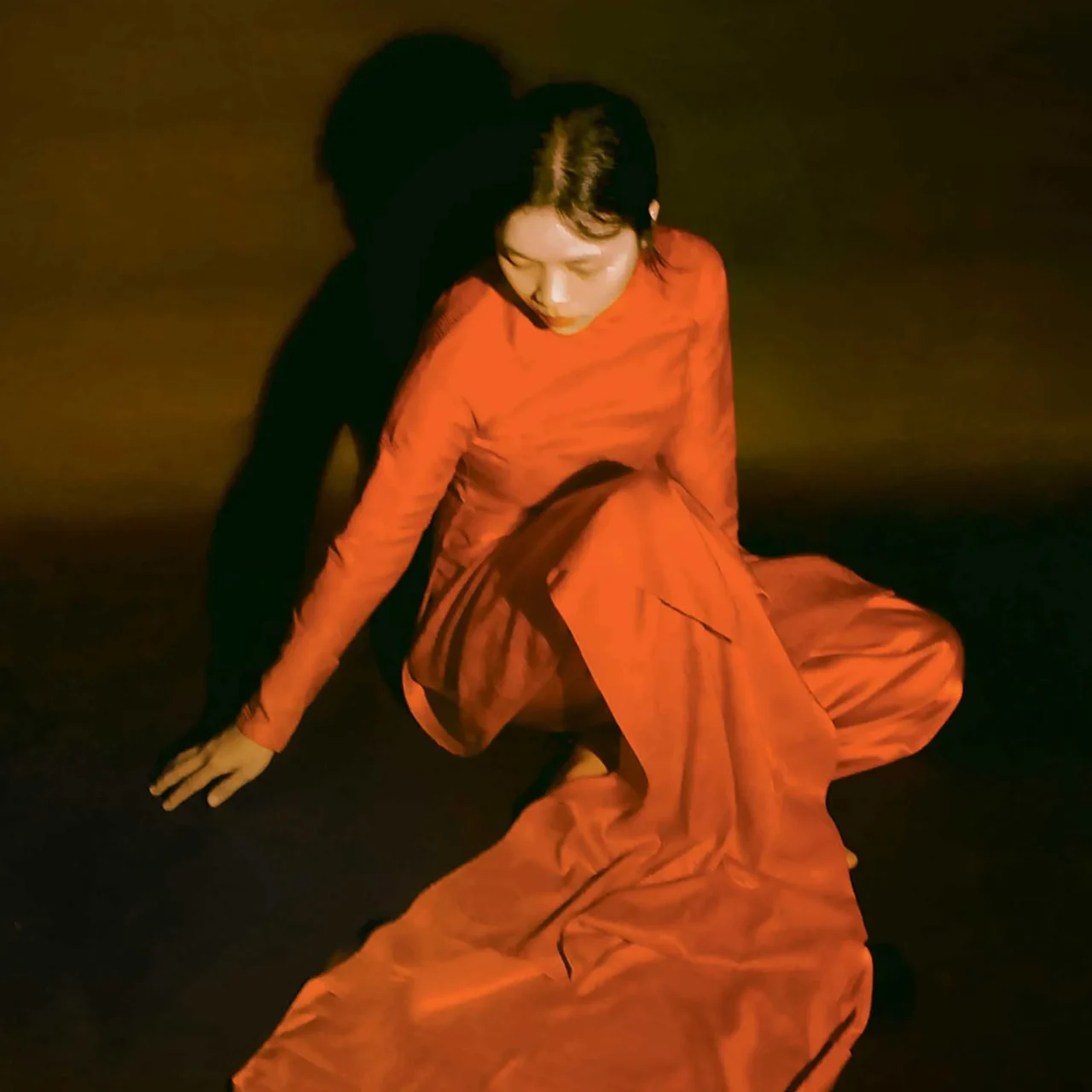 Person in a flowing orange dress crouches on a dark stage bathed in warm orange light, performing a dance pose.