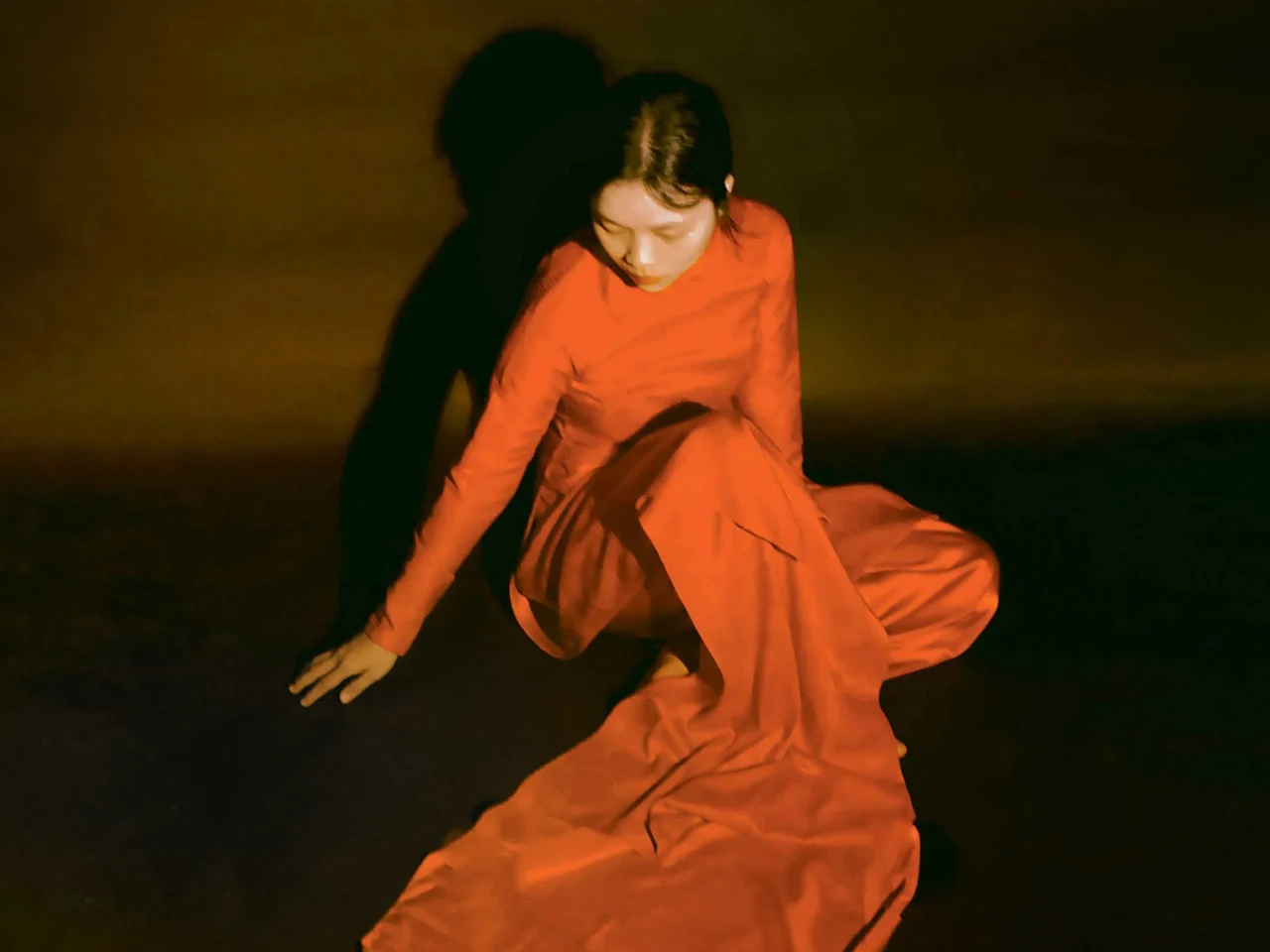 Person in a flowing orange dress crouches on a dark stage bathed in warm orange light, performing a dance pose.