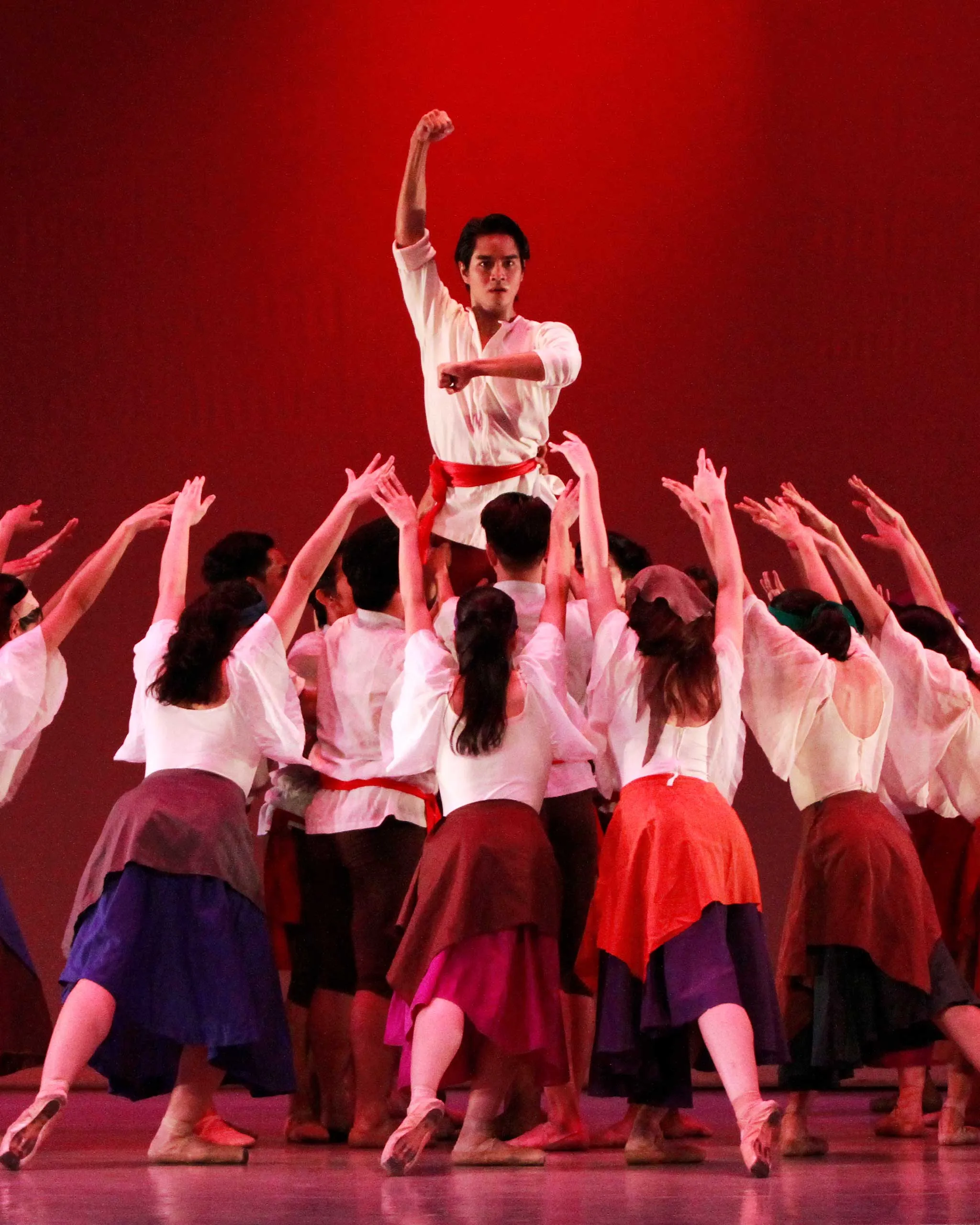 Center-stage male dancer in a white shirt with a red belt is raised above a crowd, one arm raised in a fist, surrounded by dancers in white tops and colorful skirts against a red backdrop.
