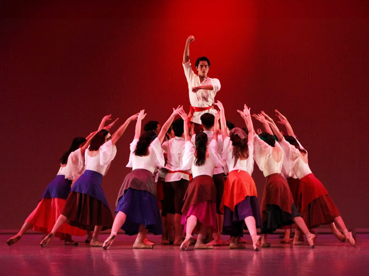 Center-stage dancer raises a clenched fist while a circle of performers in white tops and colorful skirts reach upward in a coordinated group dance against a red backdrop.