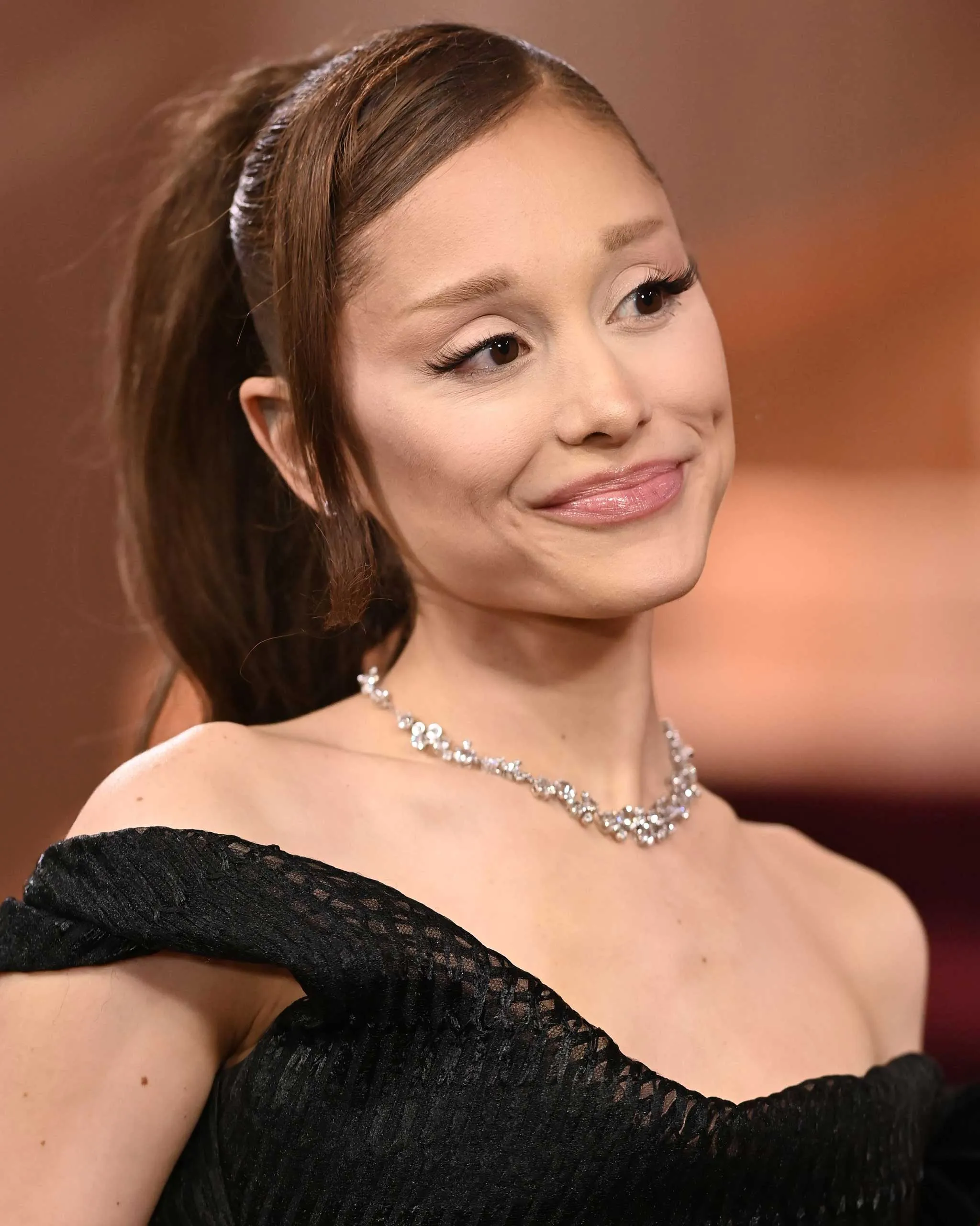 Close-up of a smiling woman wearing a black off-shoulder dress and a diamond necklace, looking to the side.