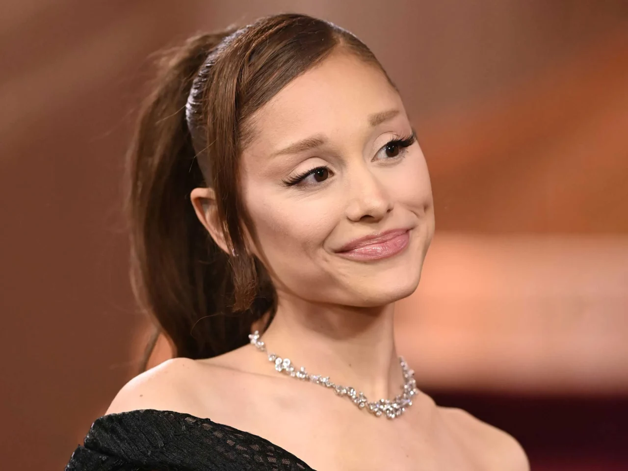 Elegant close-up of a smiling woman wearing a diamond necklace and off-shoulder black dress at a formal event, looking to the side.