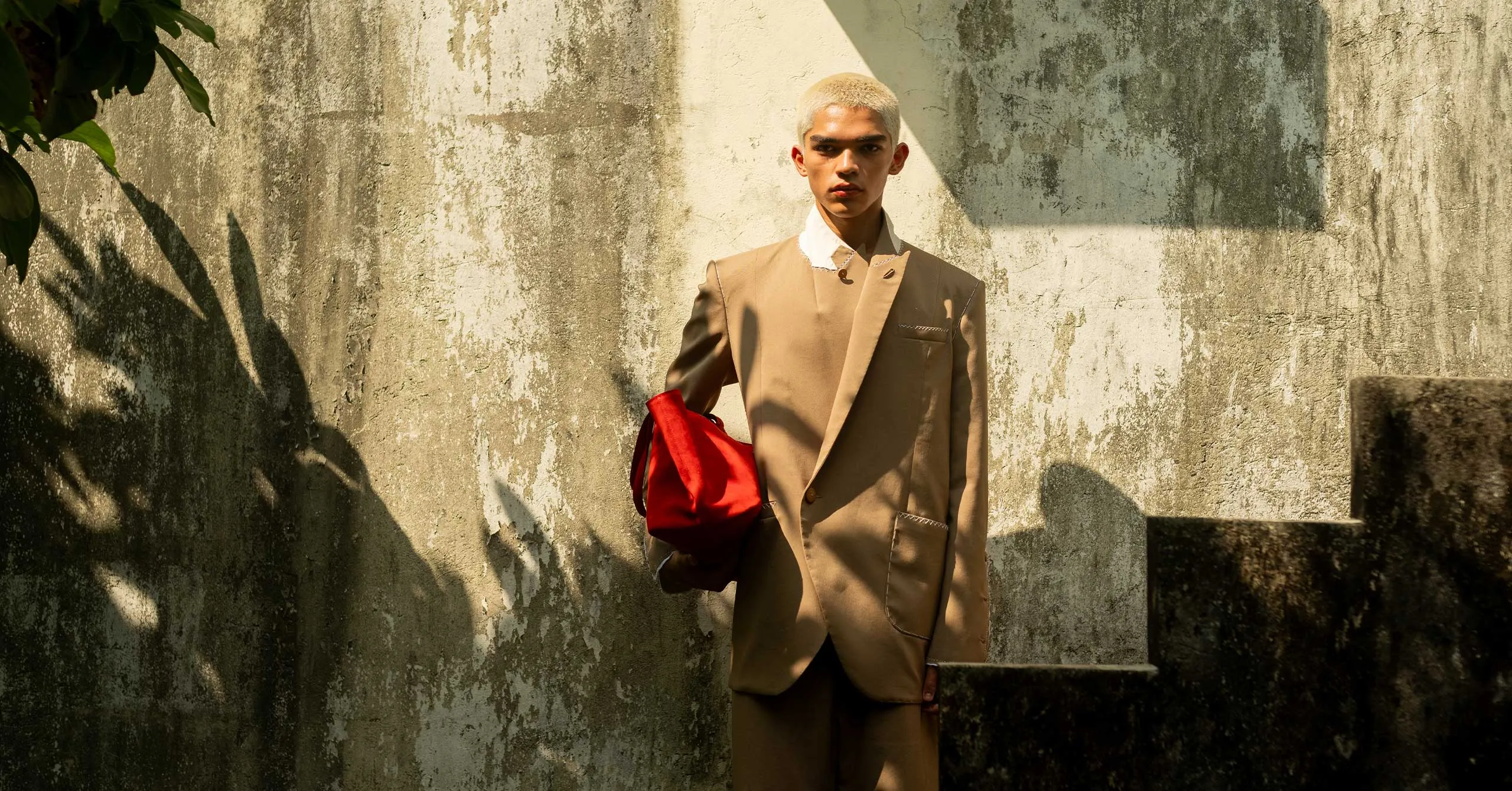 Young man with platinum blonde buzz cut wearing a beige suit, white shirt, and holding a red bag, standing against a weathered wall with shadows.