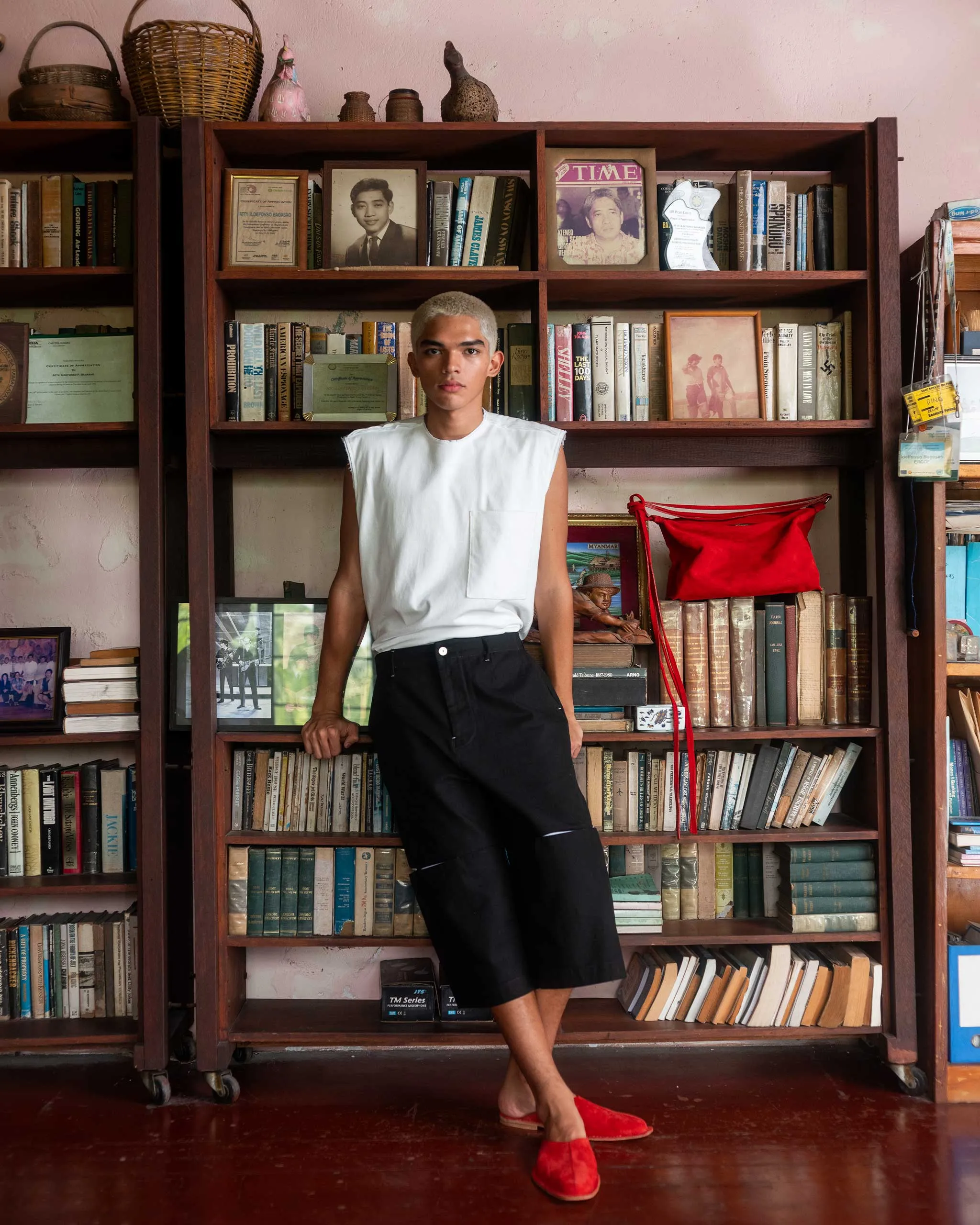 Young man with bleached hair in a white sleeveless shirt and black shorts stands in front of a tall wooden bookshelf filled with old books and framed photos, baskets on top.