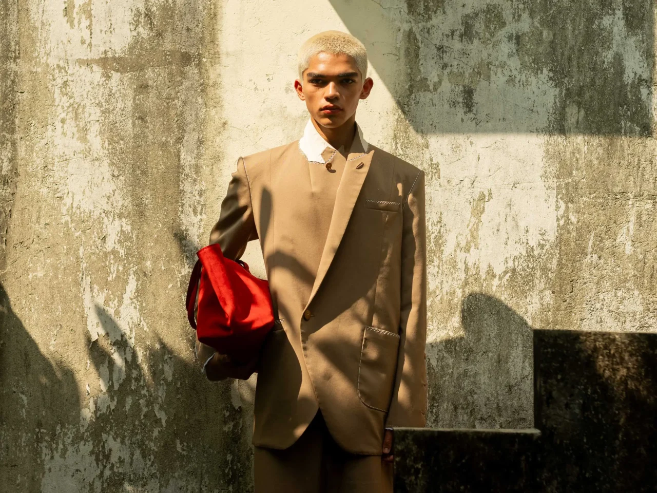 Young man with platinum blonde buzz cut wearing a beige suit, white shirt, and holding a red bag, standing against a weathered wall with shadows.