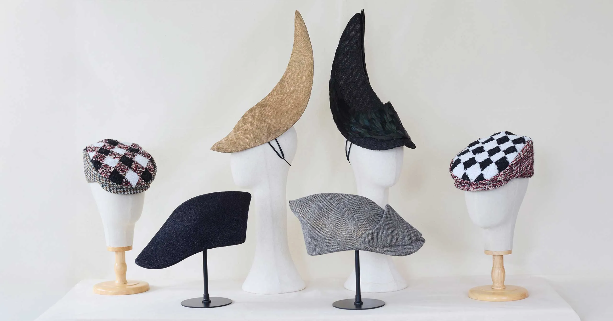 Display of fashion hats on white mannequin heads: two checkered berets (black‑white and white‑red), a black brimless cap, a gray tweed cap, and two tall pointed witch hats (beige and black).