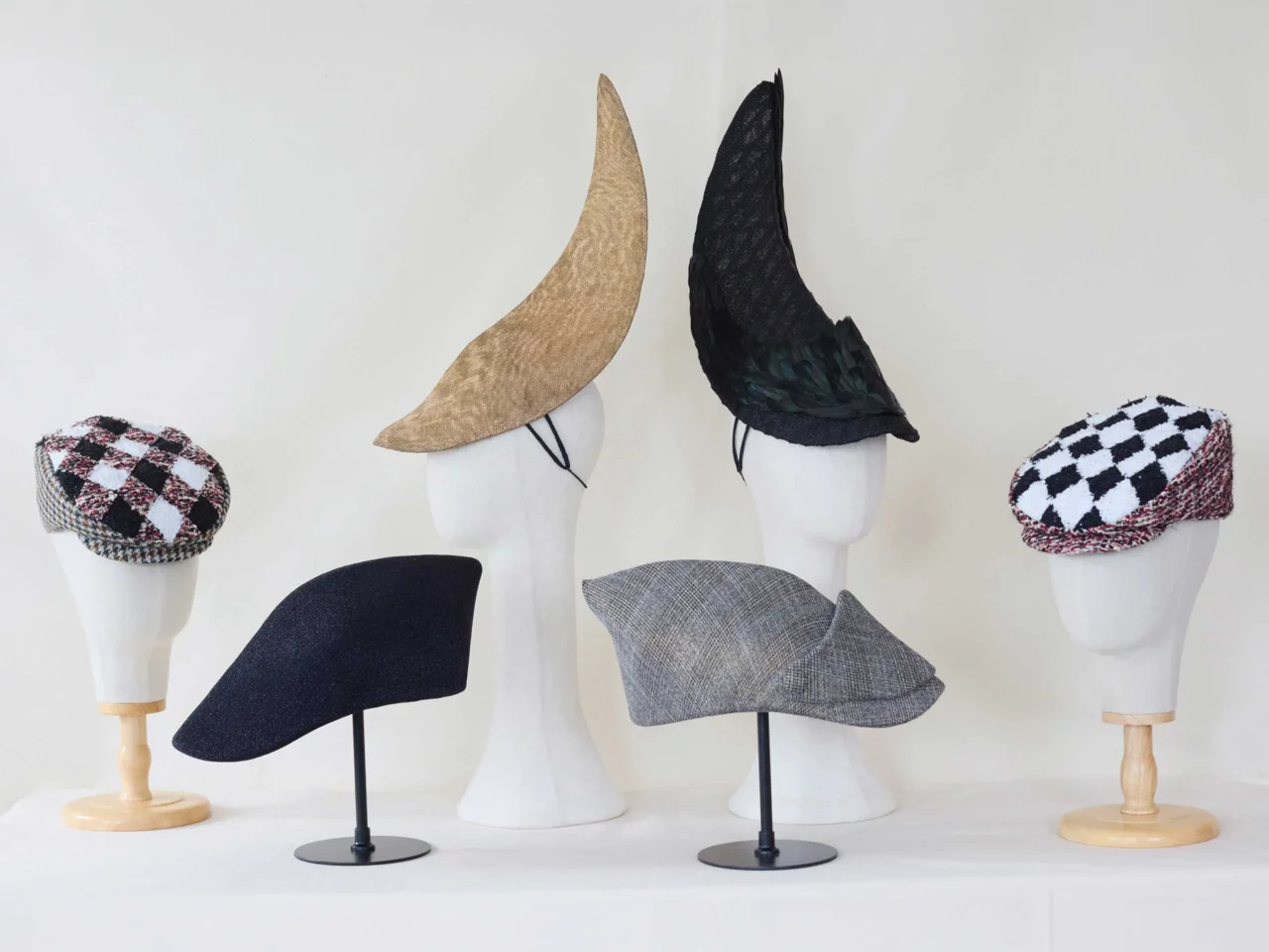 Display of fashion hats on white mannequin heads: two checkered berets (black‑white and white‑red), a black brimless cap, a gray tweed cap, and two tall pointed witch hats (beige and black).