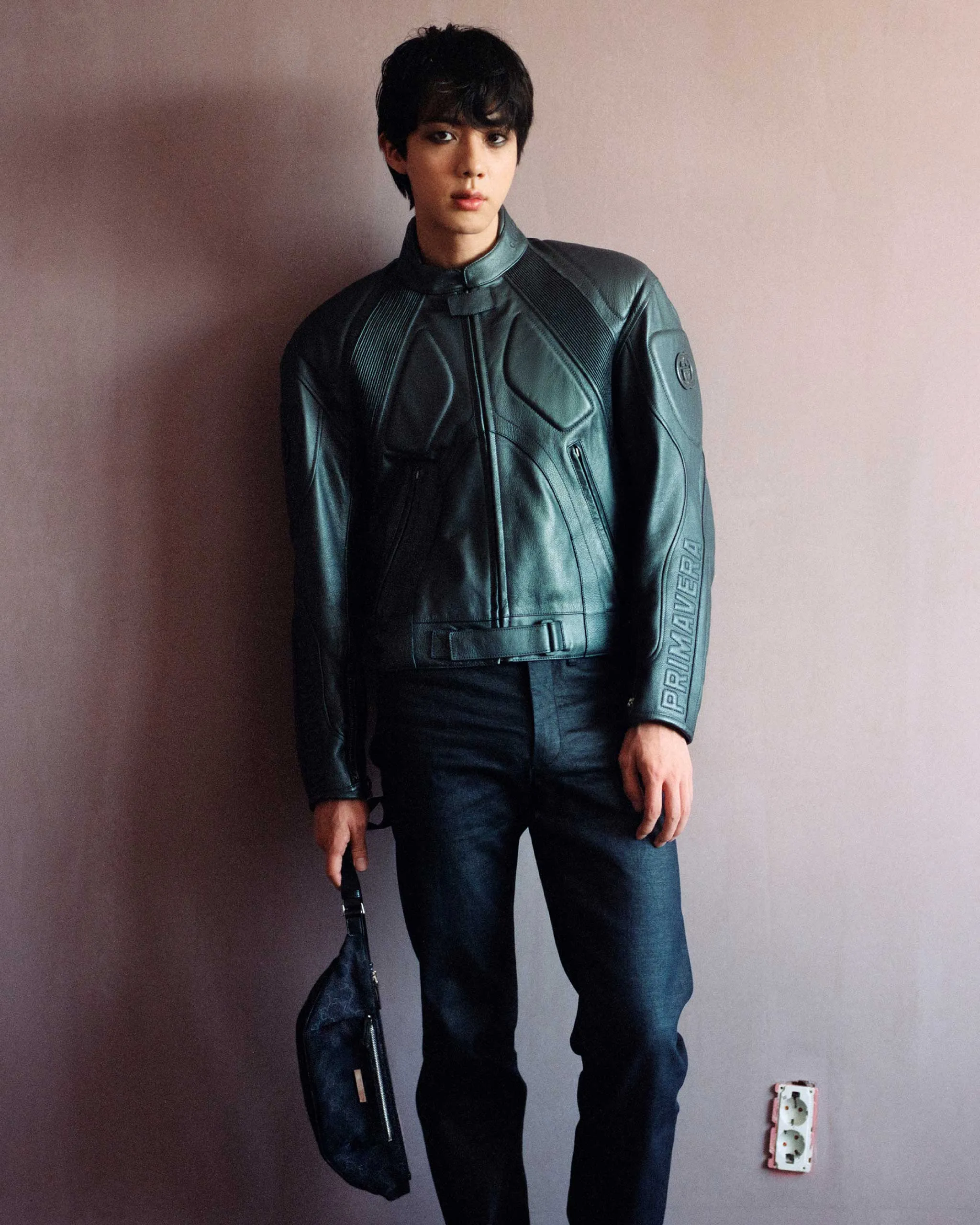 Full-body portrait of a young man in a black leather motorcycle jacket, standing against a pink wall and holding a dark bag.