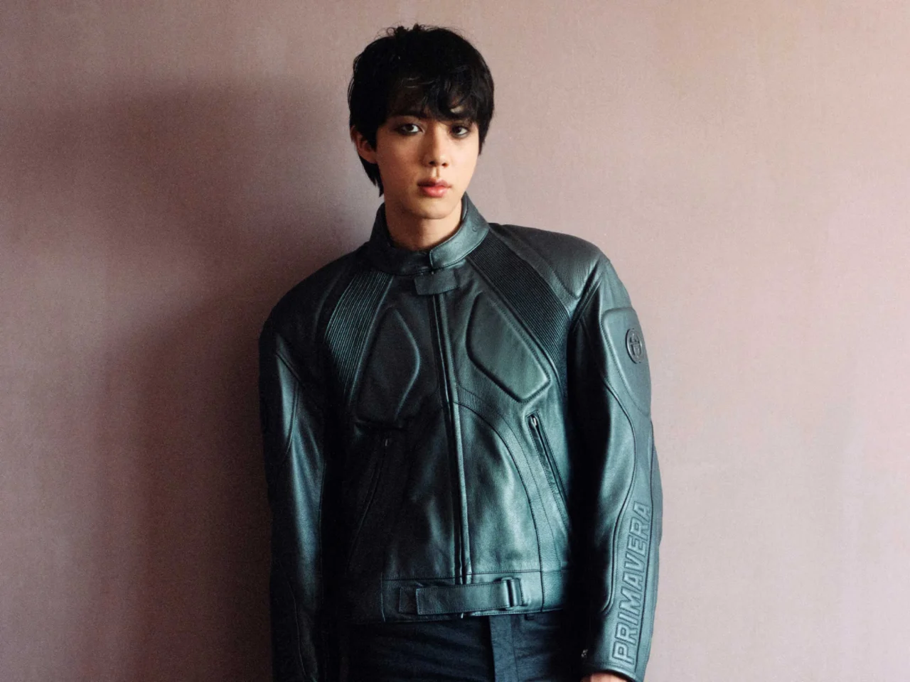 Young man with short black hair wearing a black leather jacket, standing against a beige wall.
