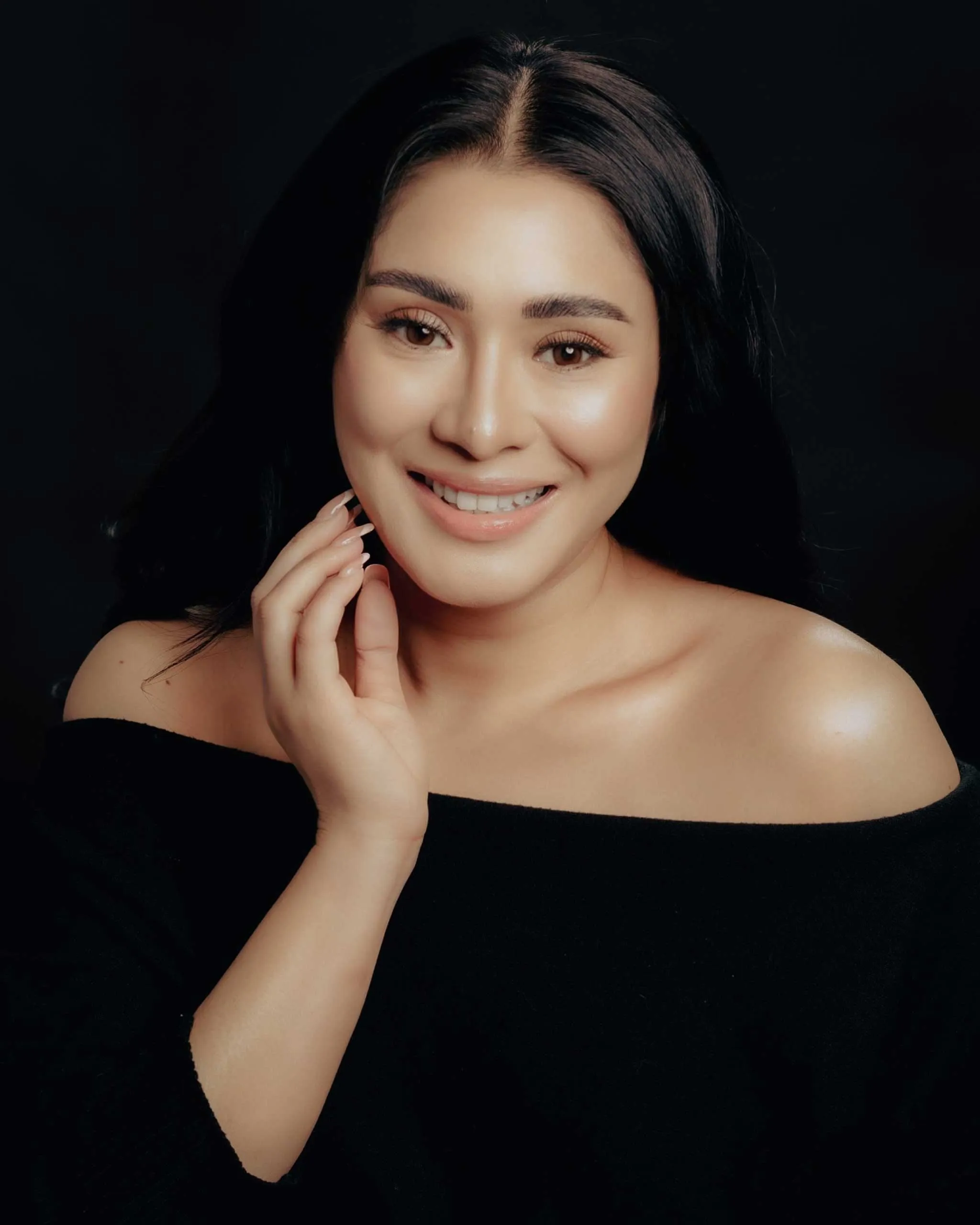 Close-up portrait of a smiling woman with dark hair and an off-the-shoulder black top against a black background, hand near her face.
