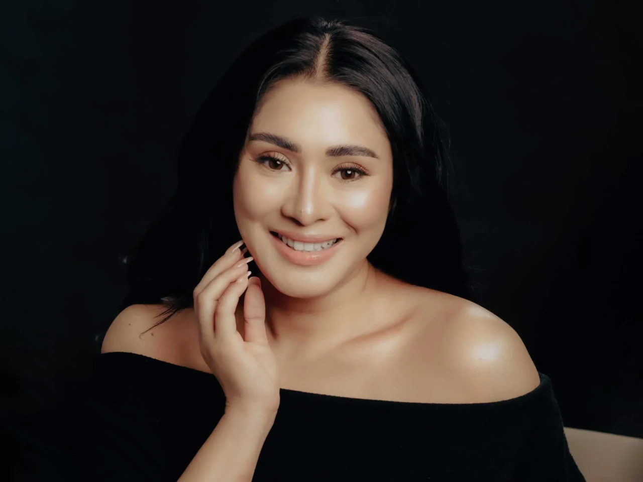 Smiling woman with dark hair in an off-shoulder black top against a dark background, hand near chin.