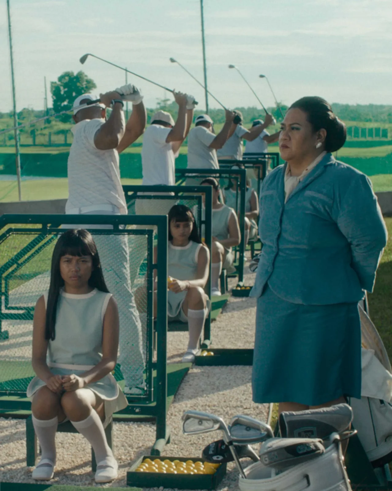 People in white outfits line up with golf clubs at a driving range, while a woman in a blue suit watches on the right.