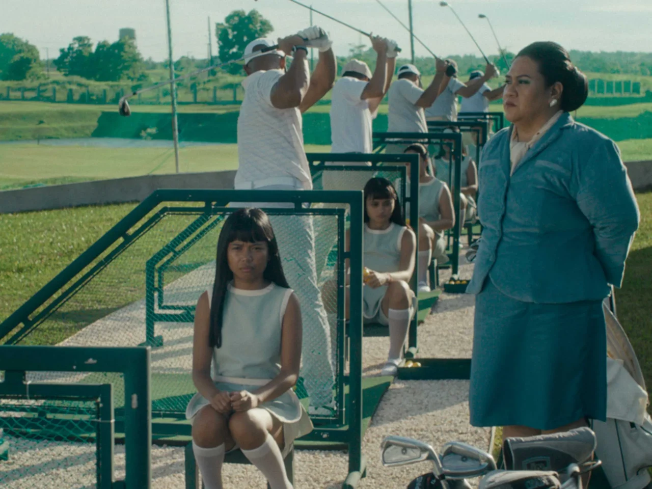 People in white outfits line up with golf clubs at a driving range, while a woman in a blue suit watches on the right.