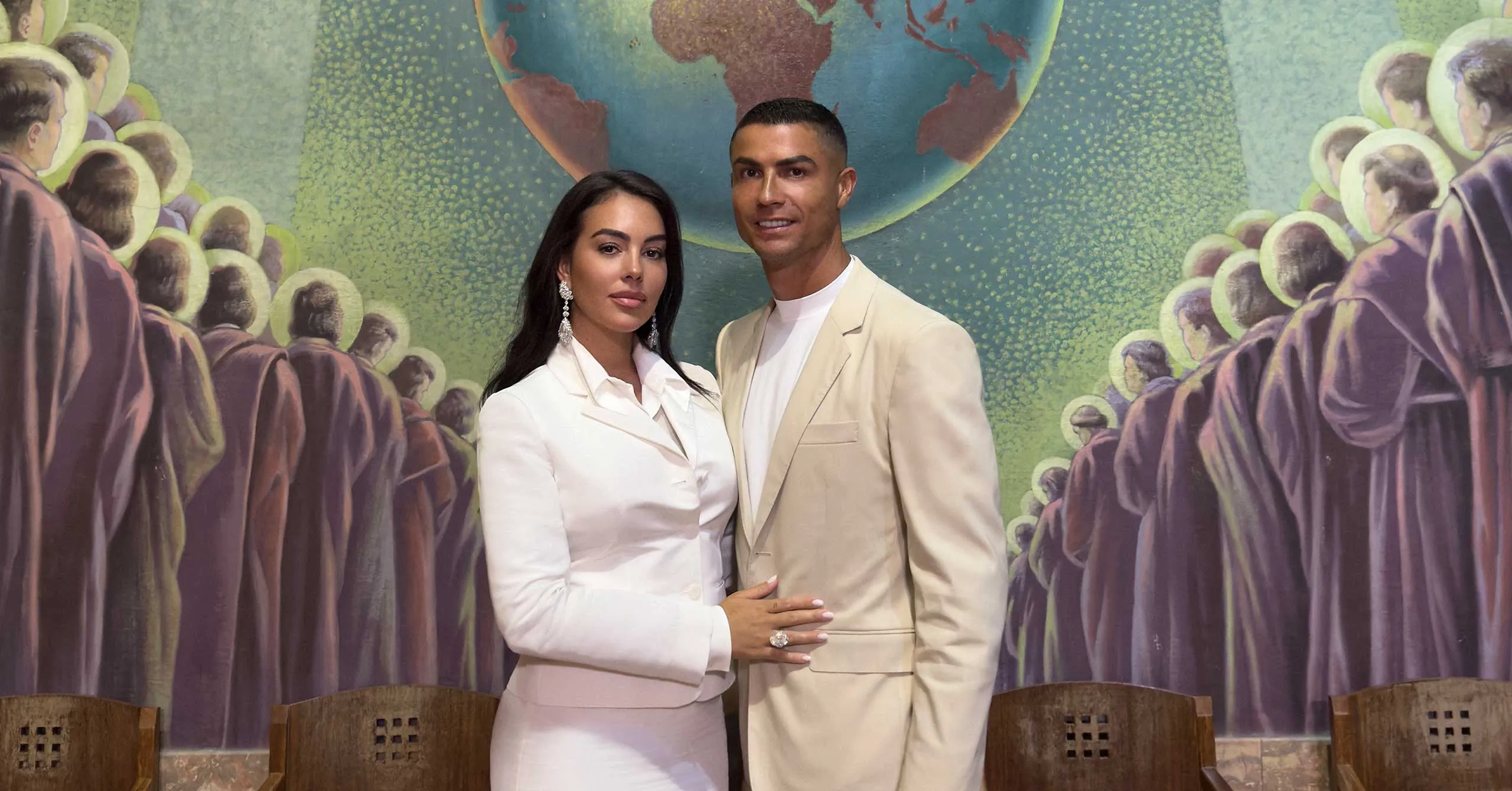 Couple posing together in light-colored formal outfits against a colorful mural featuring a globe and crowd silhouettes.