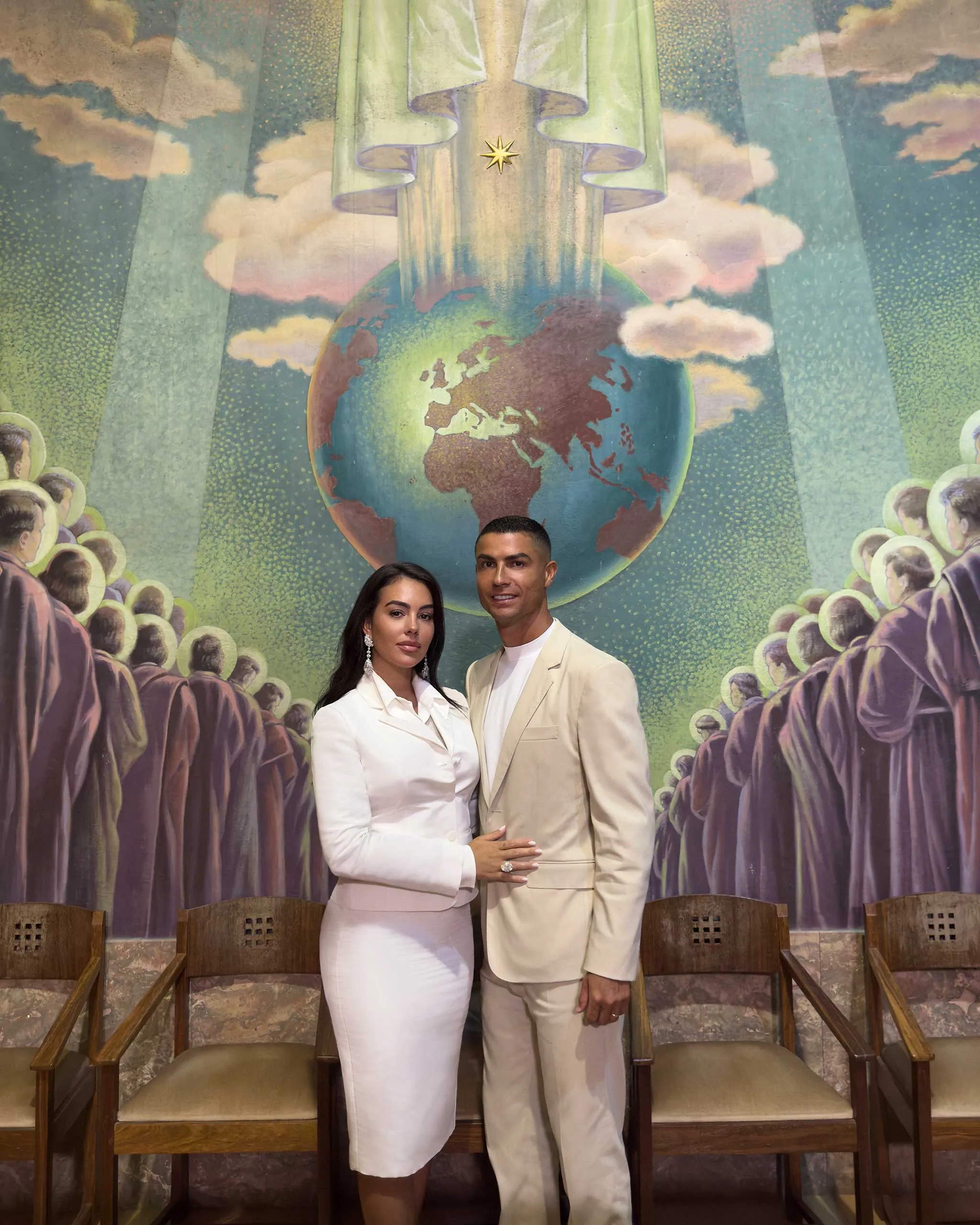 Two people in white and beige formal outfits pose in front of a colorful mural showing Earth and a crowd around it.