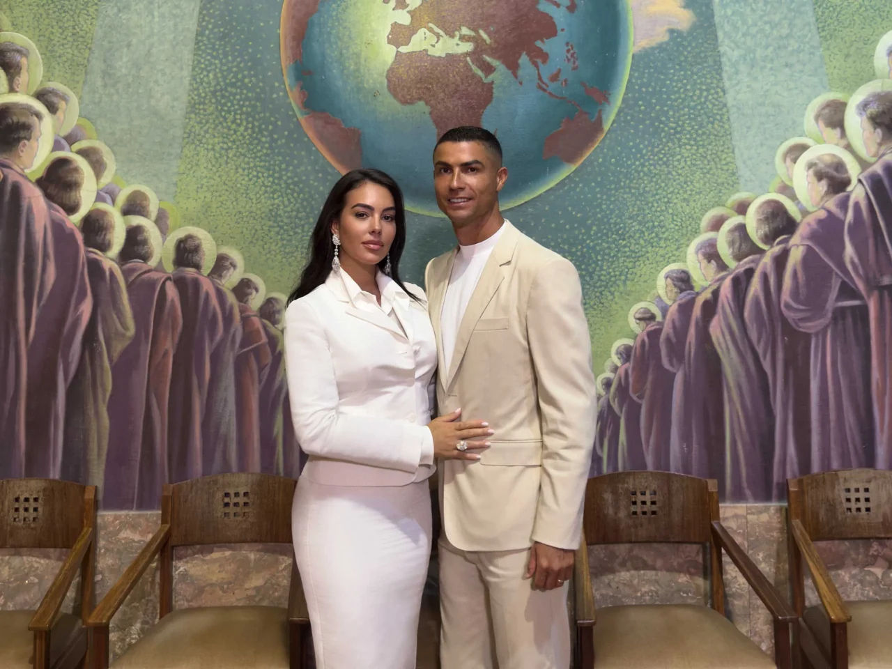 Couple posing together in light-colored formal outfits against a colorful mural featuring a globe and crowd silhouettes.