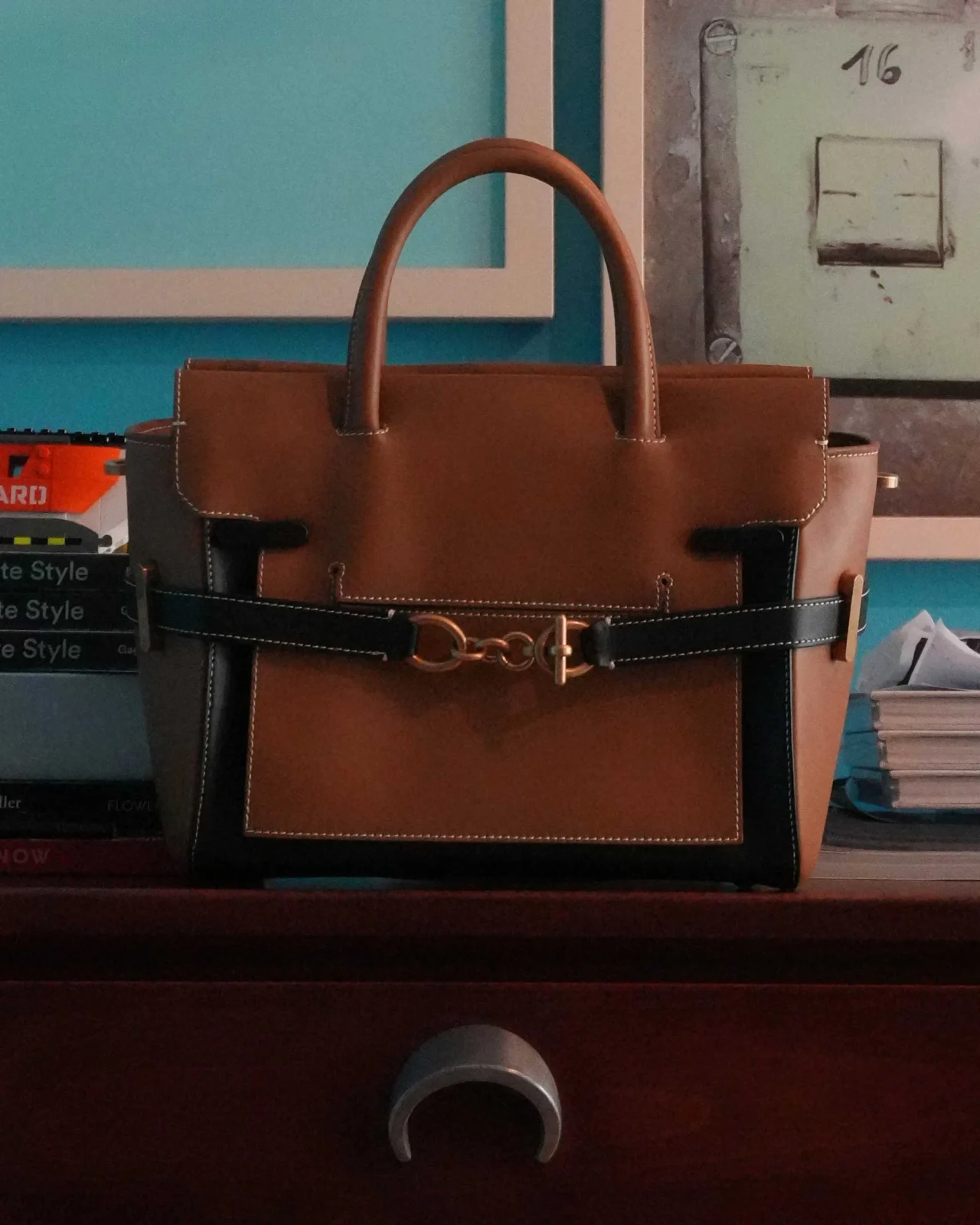 Leather handbags on a display rack in tan, black, and blue with double handles and white stitching, showcasing front pockets and gold-toned hardware.