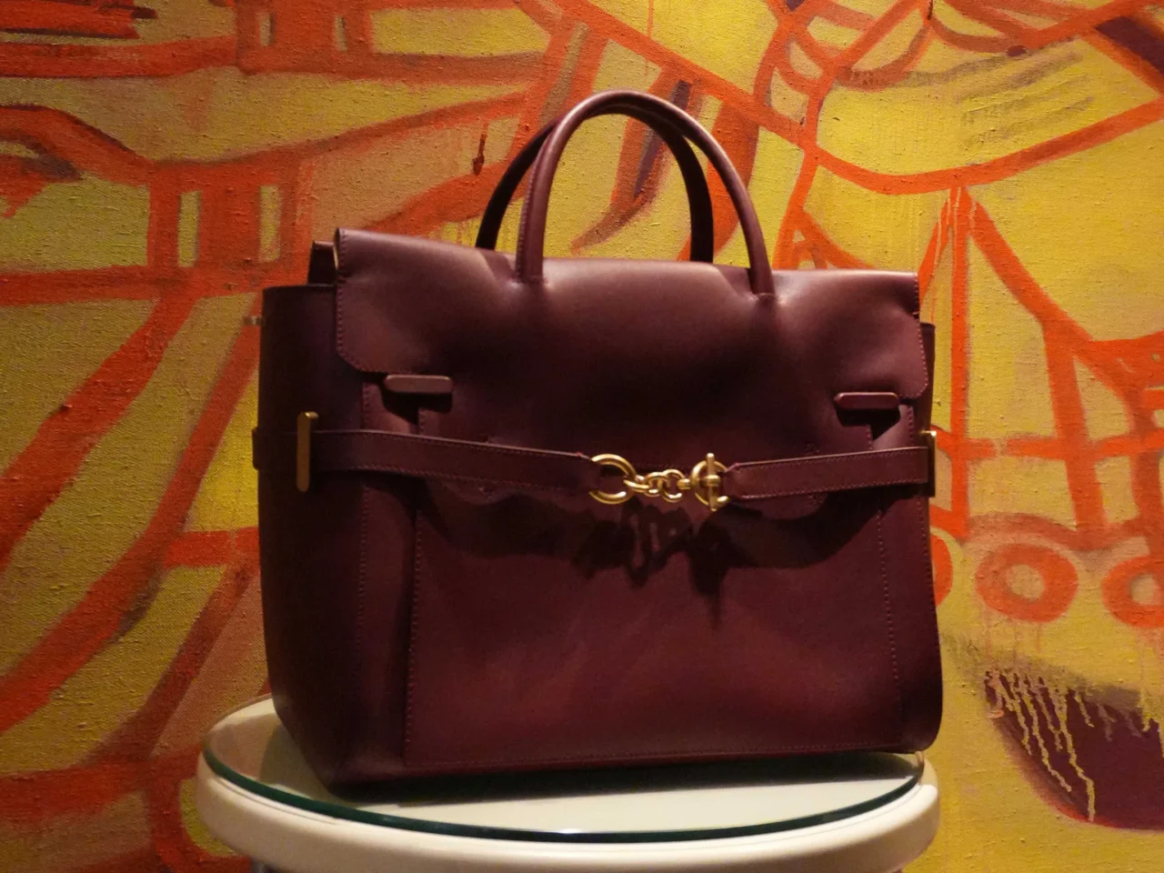 Leather handbags on a display rack in tan, black, and blue with double handles and white stitching, showcasing front pockets and gold-toned hardware.