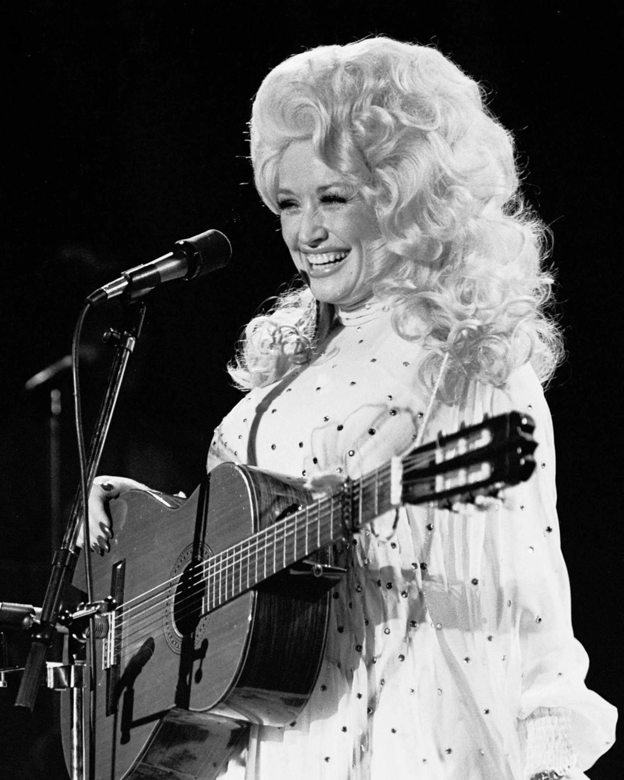 Female singer wearing a light dotted dress, smiling at a microphone while playing an acoustic guitar on stage (black and white)