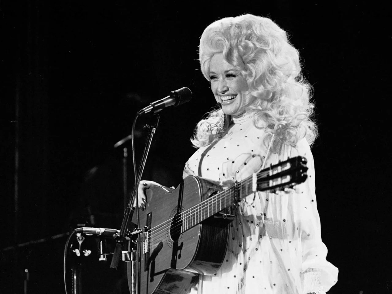 Black-and-white photo of a smiling female musician singing into a microphone while strumming an acoustic guitar on stage.