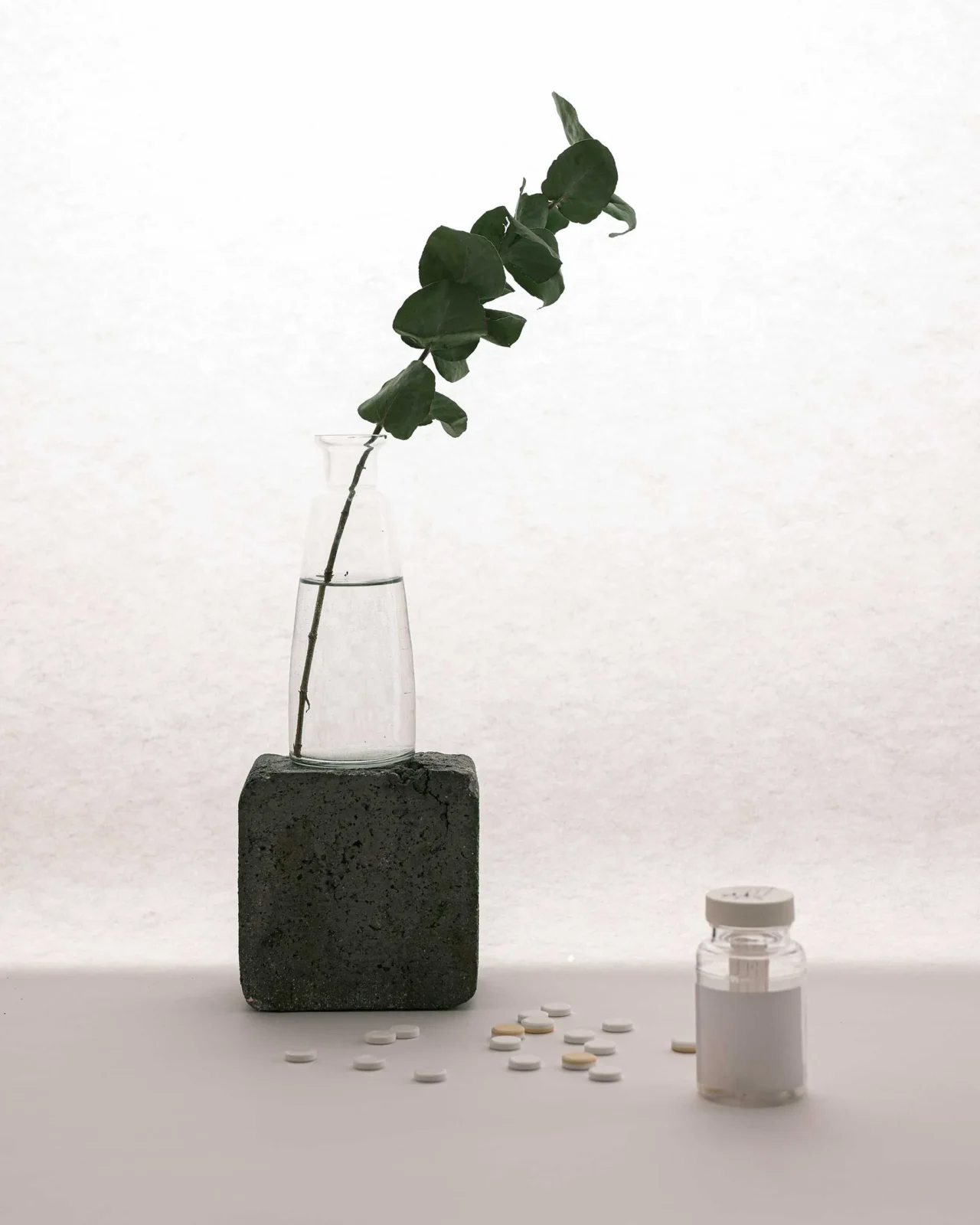 Assorted white and beige tablets scattered on a white surface next to a small glass bottle and a dark brick-shaped block on the left.